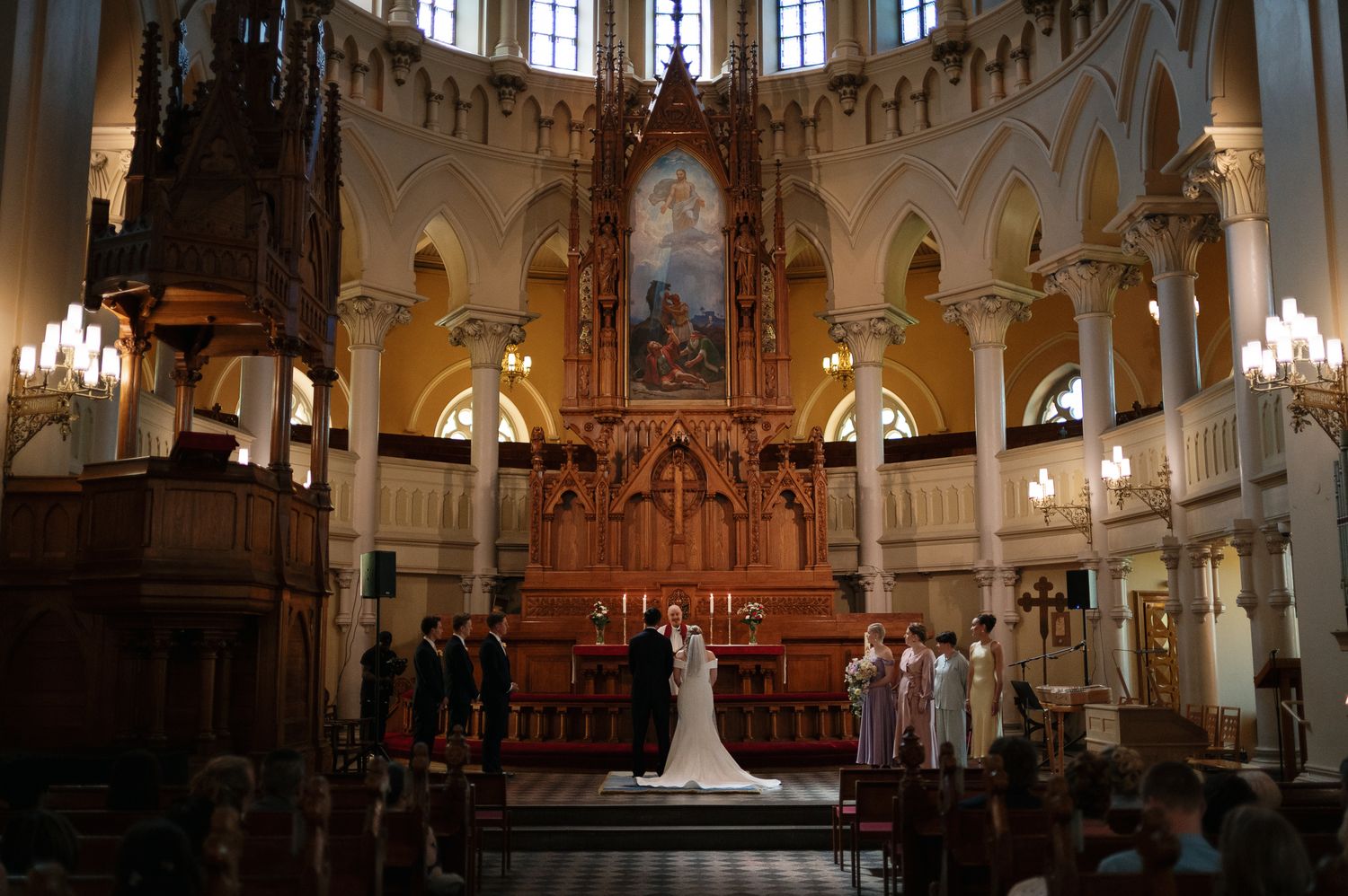 Elegant wedding ceremony at ornate church altar with bride and groom surrounded by wedding party under grand architecture.