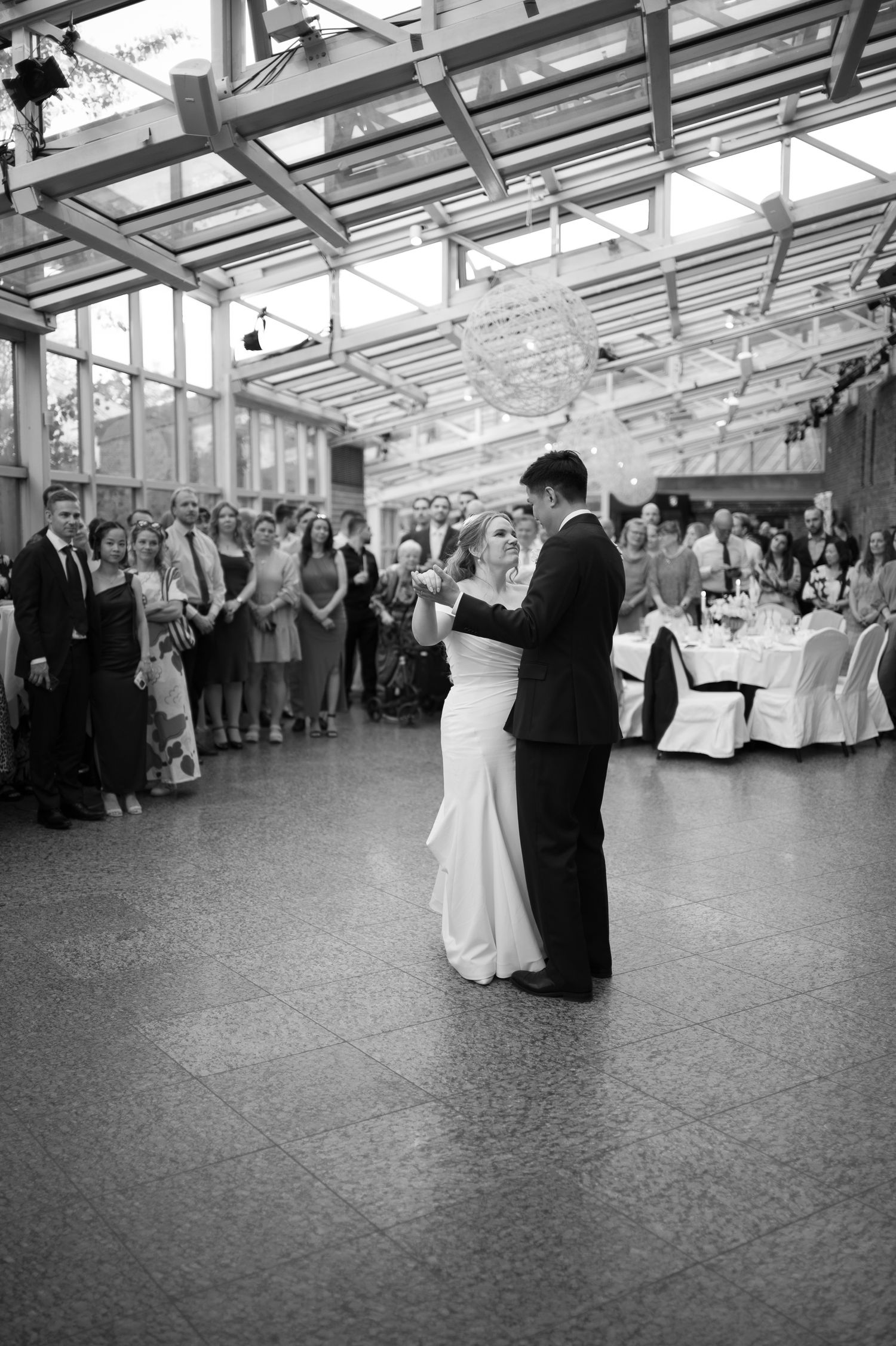Black and white photo of a bride and groom sharing their first dance at a wedding reception with guests watching.