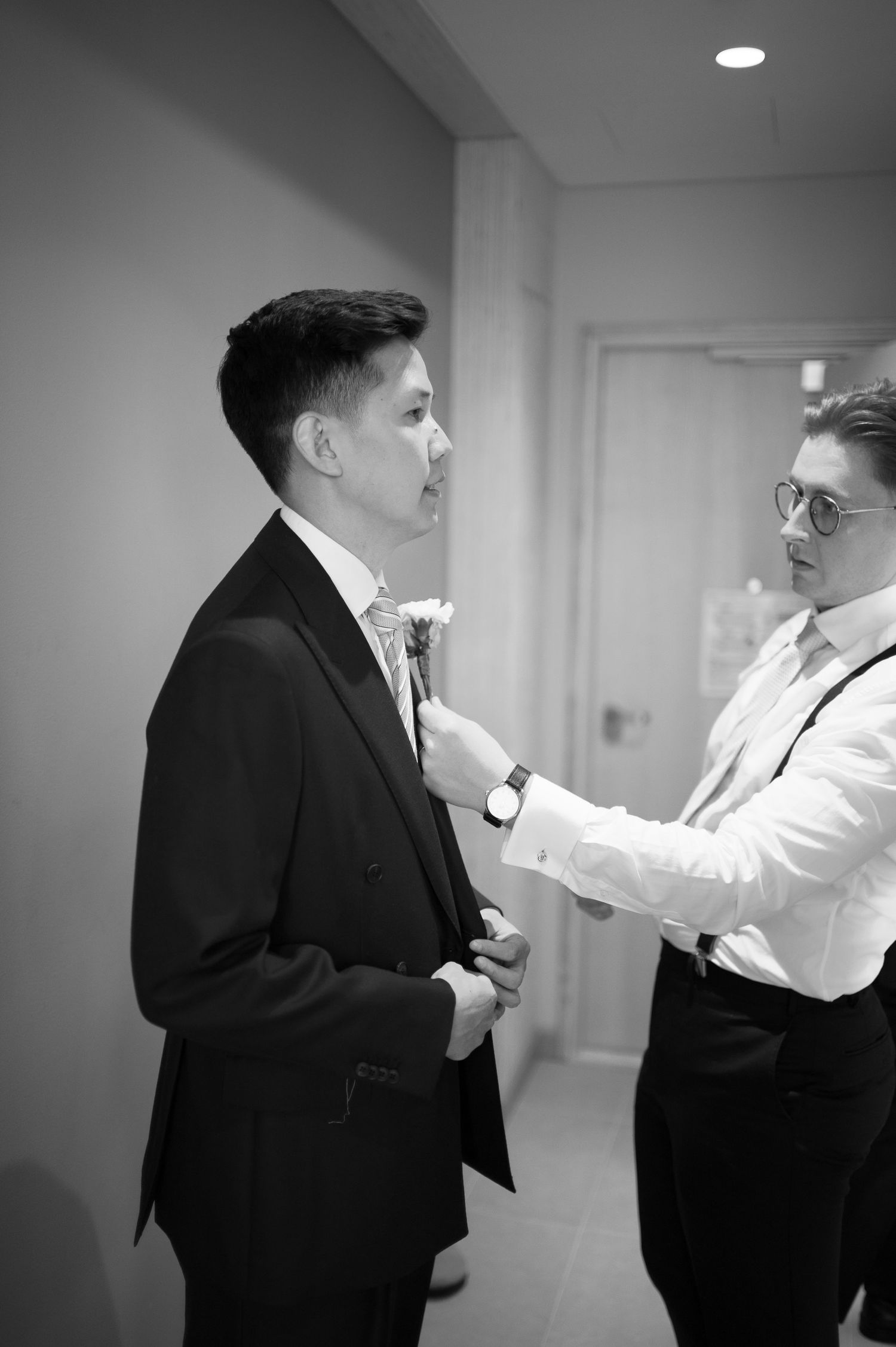 Black and white photo of a man in a suit having a boutonniere pinned to his lapel by another man in formal attire.