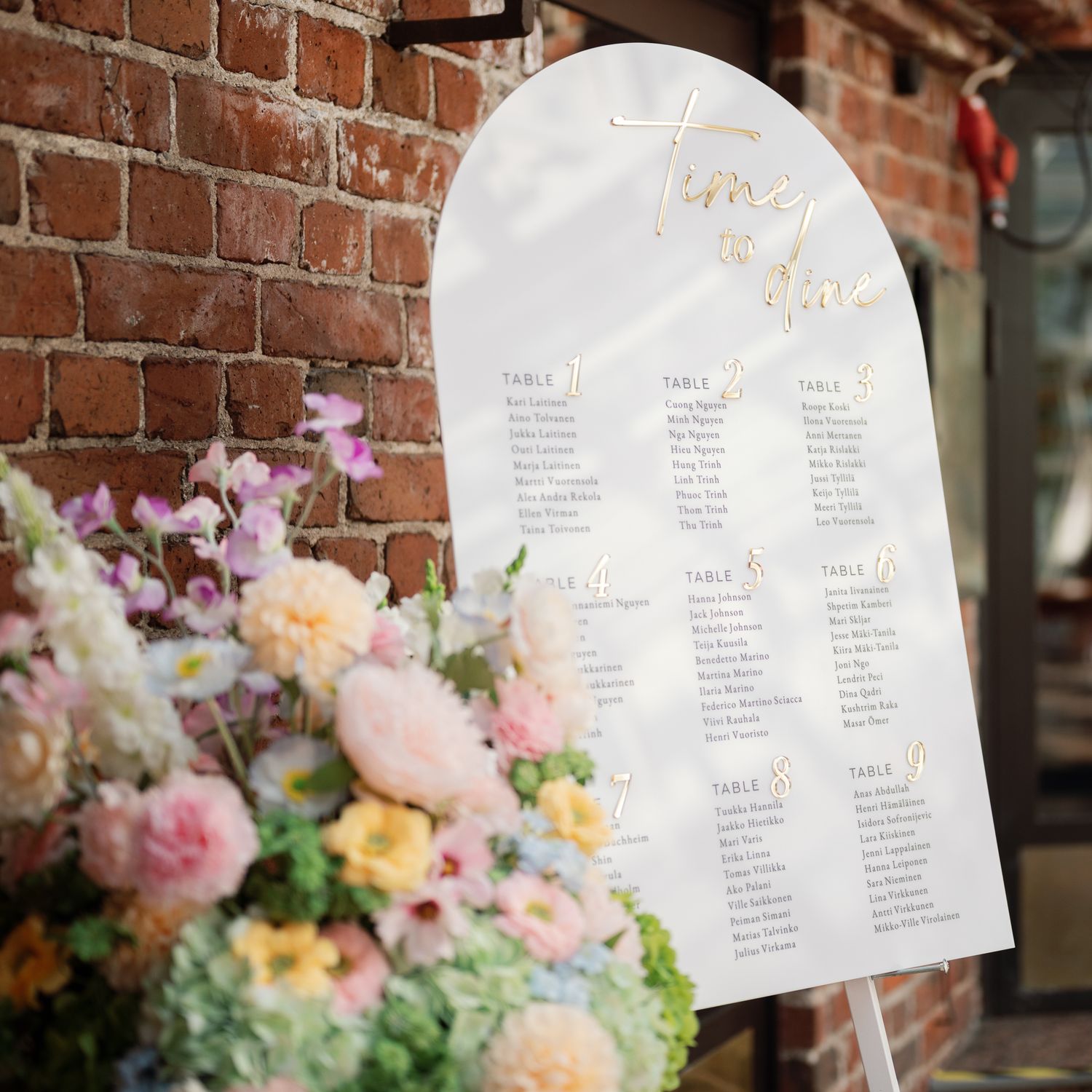 Elegant wedding seating chart on white arch sign with gold script, displayed on easel beside colorful floral arrangement.