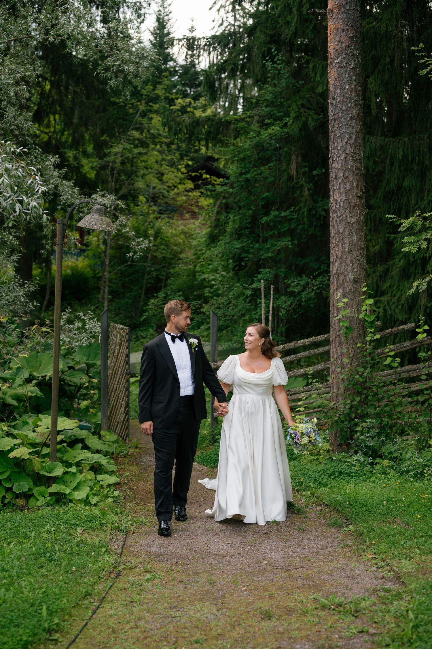 A bride and groom walking together on a forest trail surrounded by lush greenery.