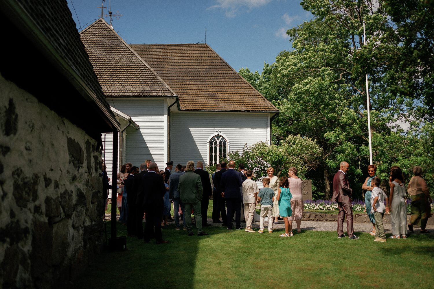 Guests gather outside a charming white wooden church surrounded by lush greenery on a sunny day.
