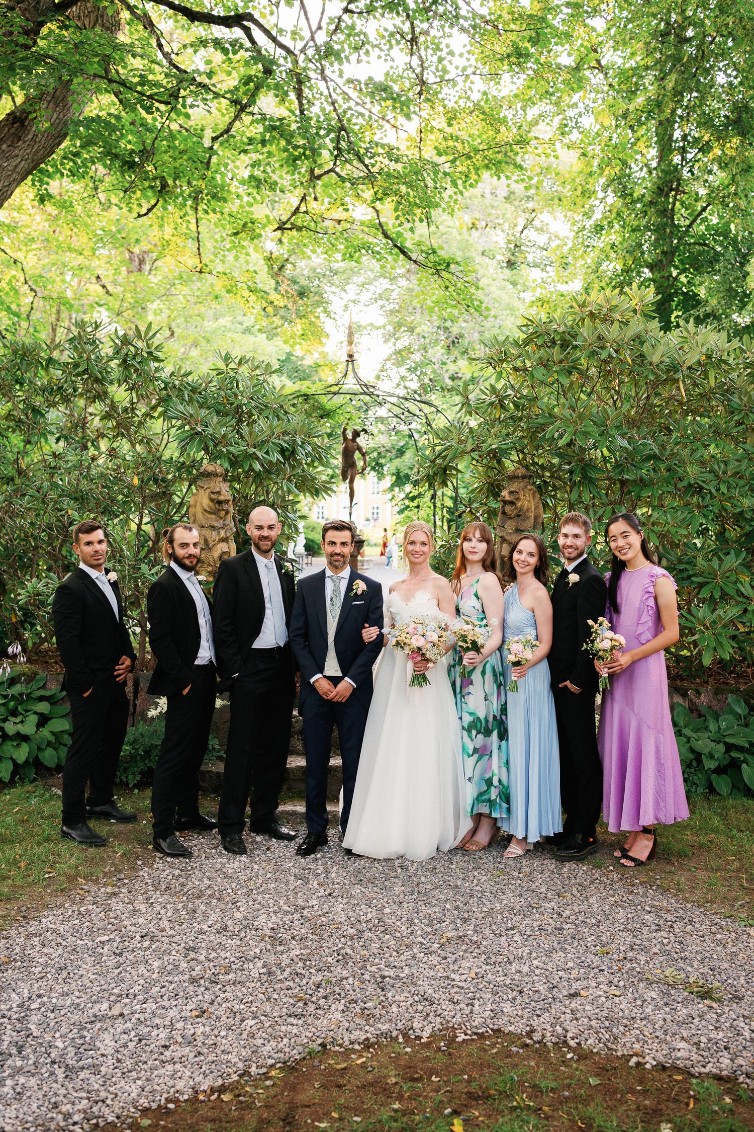 Wedding party poses in lush garden, bride in white gown with bouquet, groomsmen in suits, bridesmaids in pastel dresses.