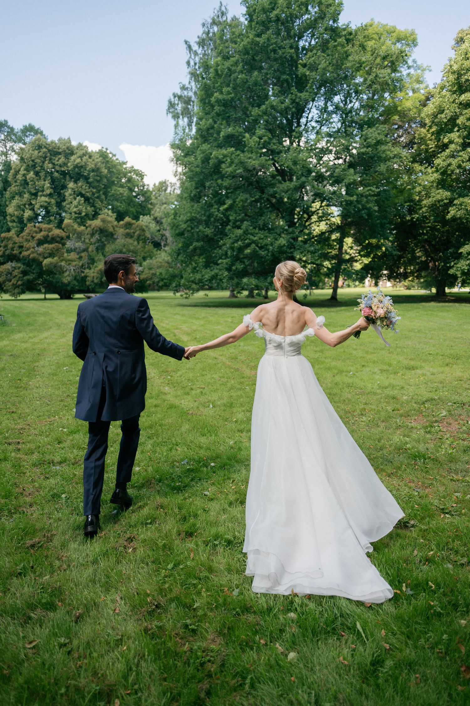 Bride and groom holding hands walking through lush green park, bride in white off-shoulder gown holding bouquet.