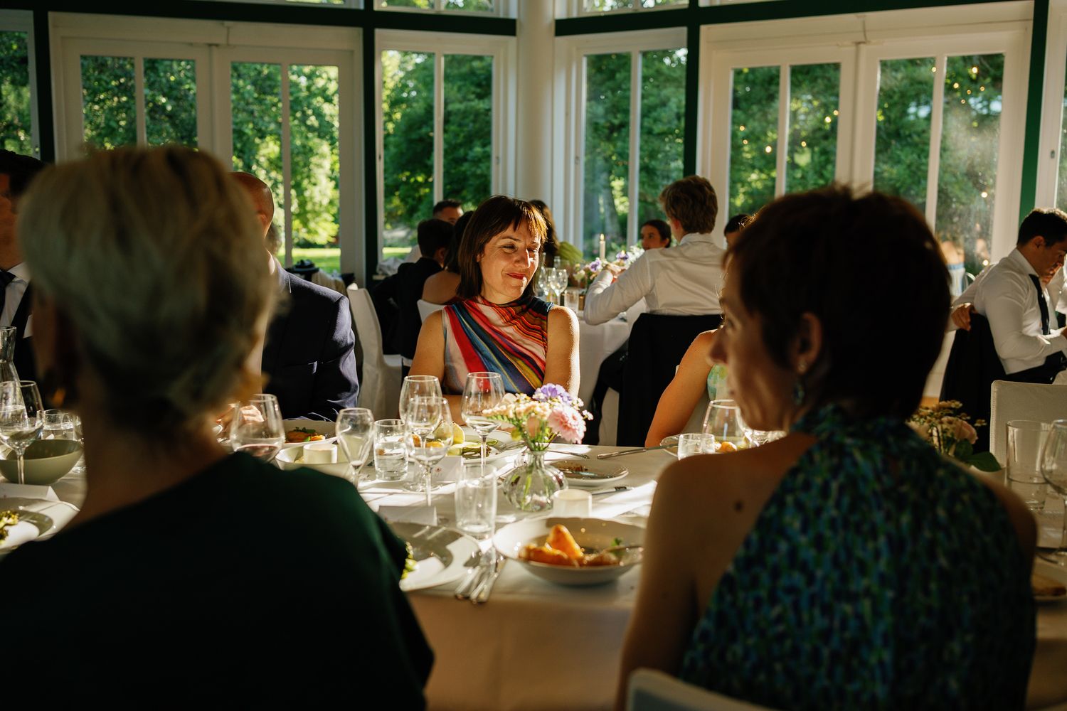 Elegant wedding reception dinner in a bright conservatory with guests in formal attire seated at beautifully set tables.