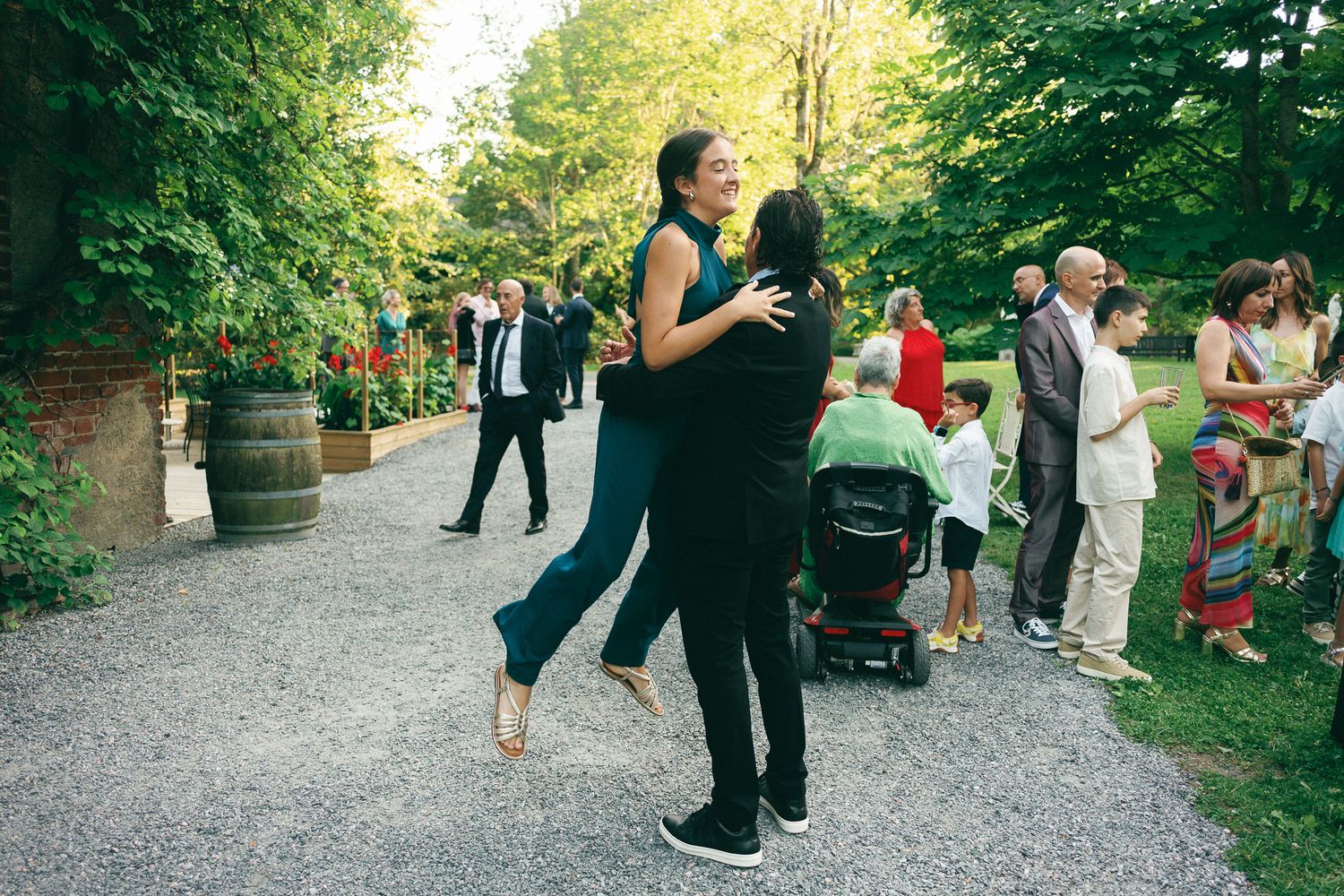 Couple dancing outdoors at a garden party with guests mingling on a gravel path surrounded by lush green trees.