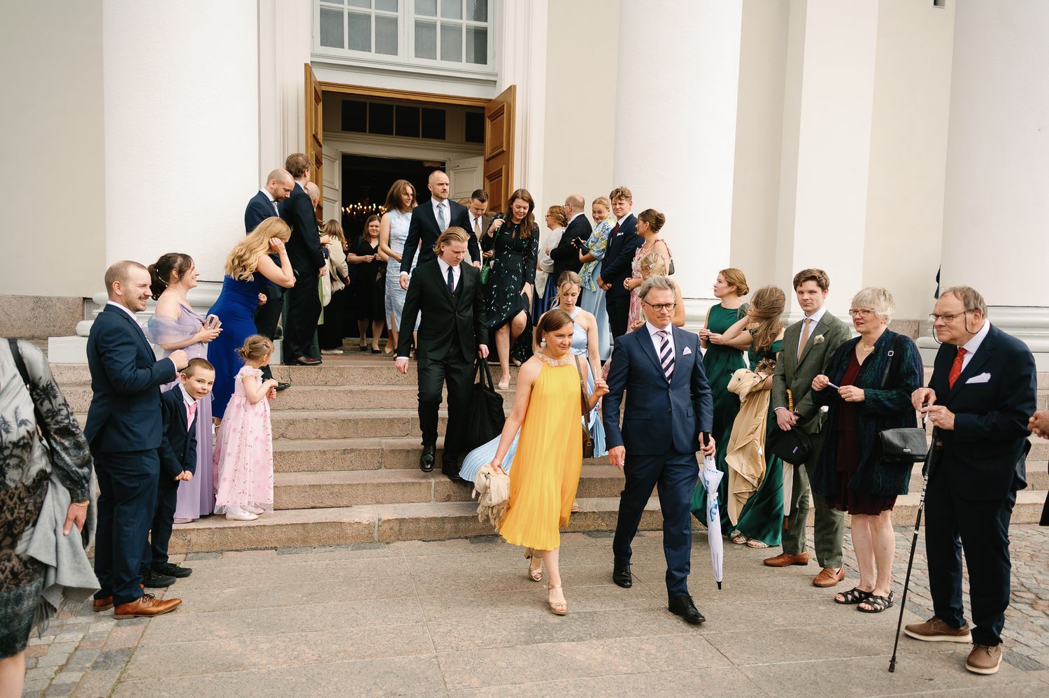 Wedding guests descend church steps in formal attire, with a woman in yellow dress leading the procession.