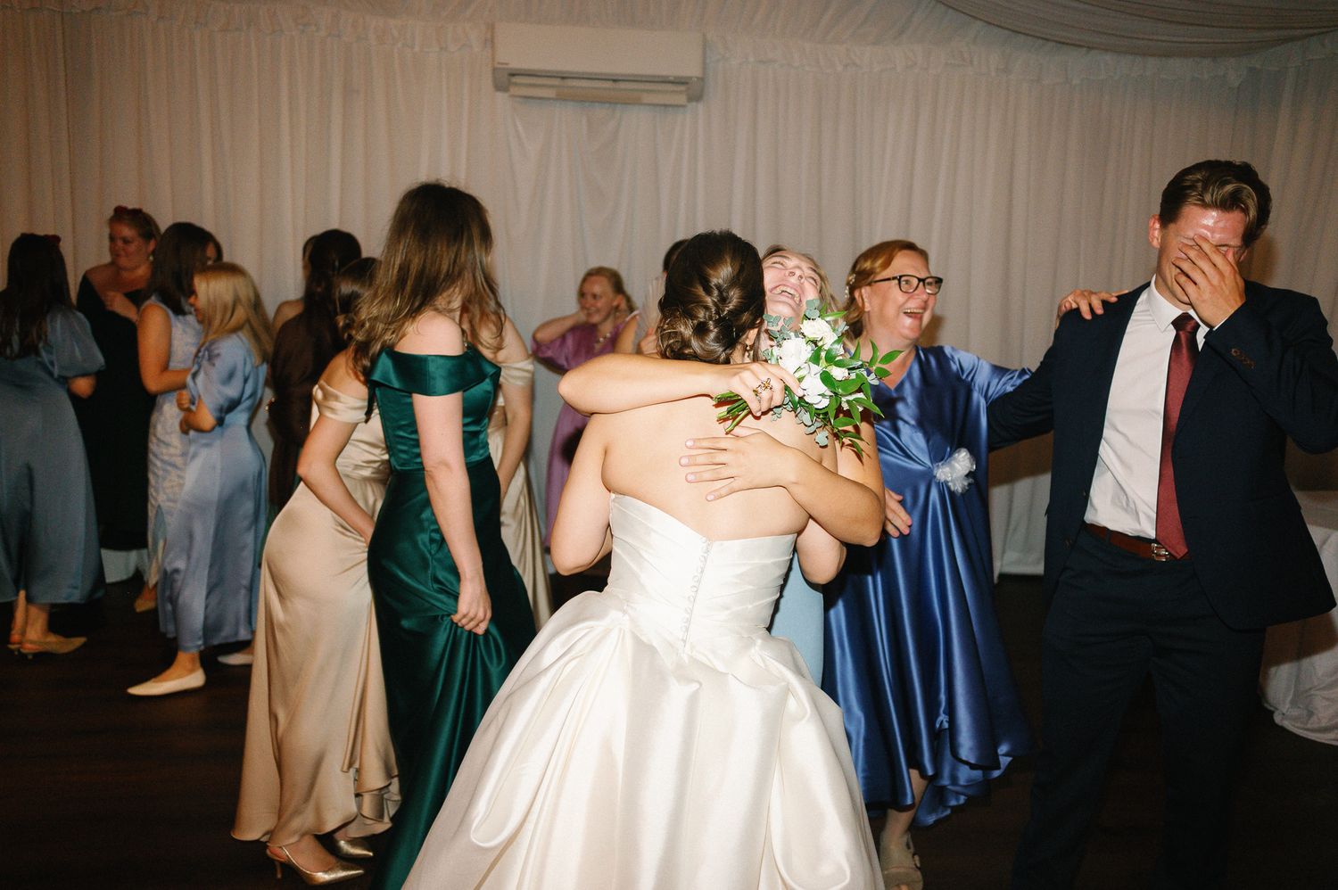 Bride in white gown hugs guest holding bouquet on wedding reception dance floor surrounded by elegantly dressed attendees.