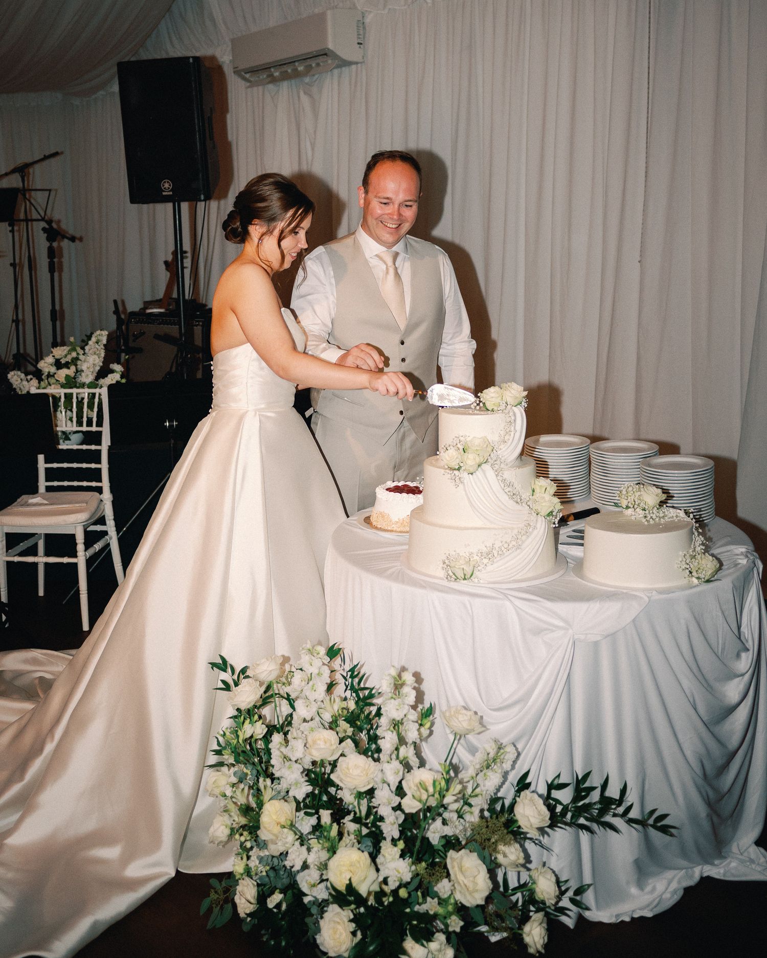 Bride and groom cutting a white tiered wedding cake surrounded by white floral arrangements at their reception.