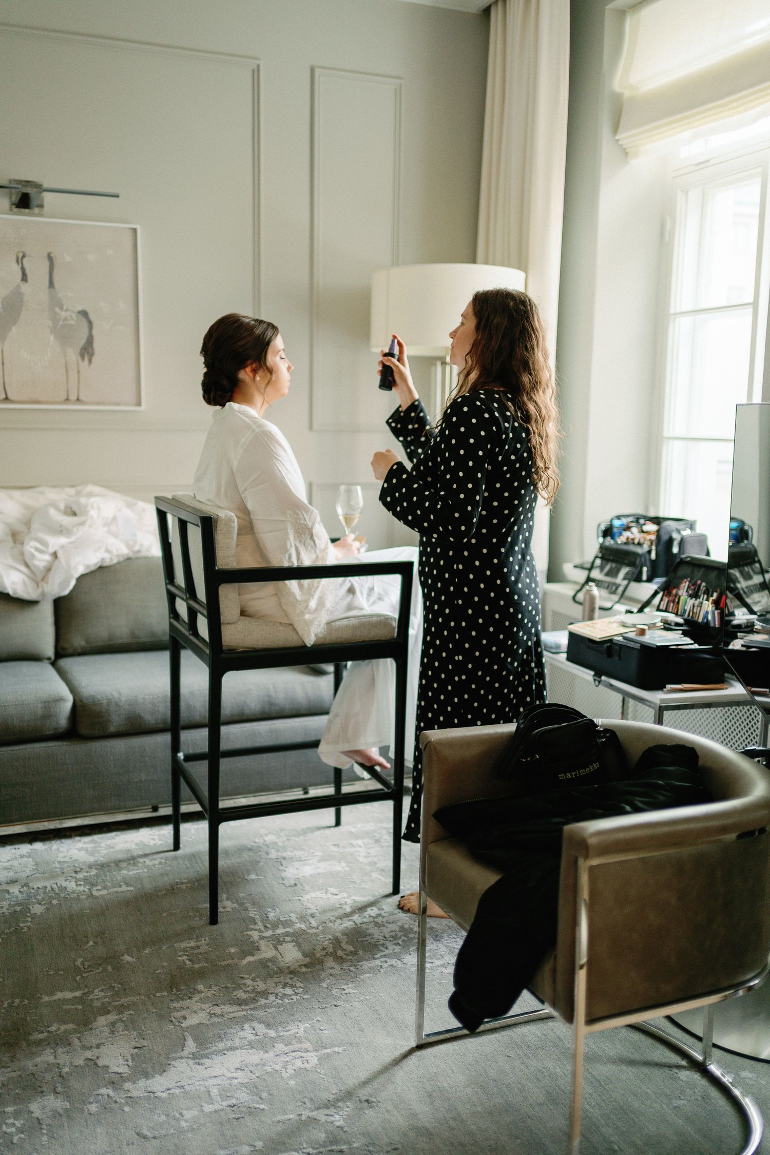 A makeup artist in a polka dot robe applies makeup to a bride in a white robe in an elegant hotel suite.