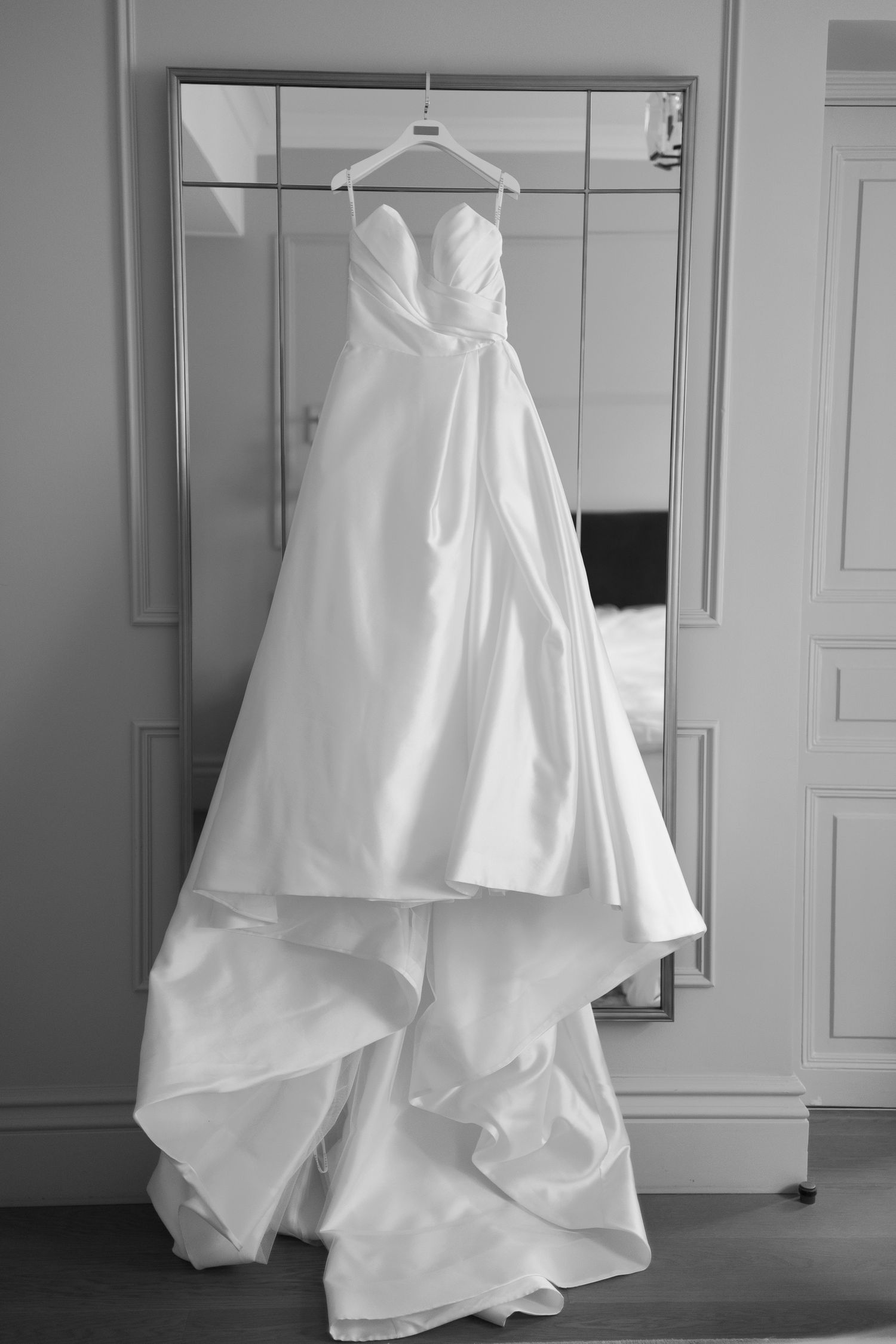 Elegant strapless white wedding dress hanging on a hanger in front of a mirror, black and white photo.