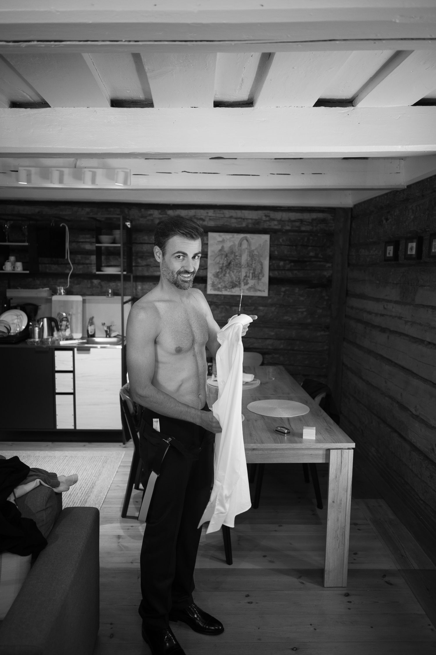 Shirtless man holding white shirt in rustic wooden cabin interior, black and white photography.