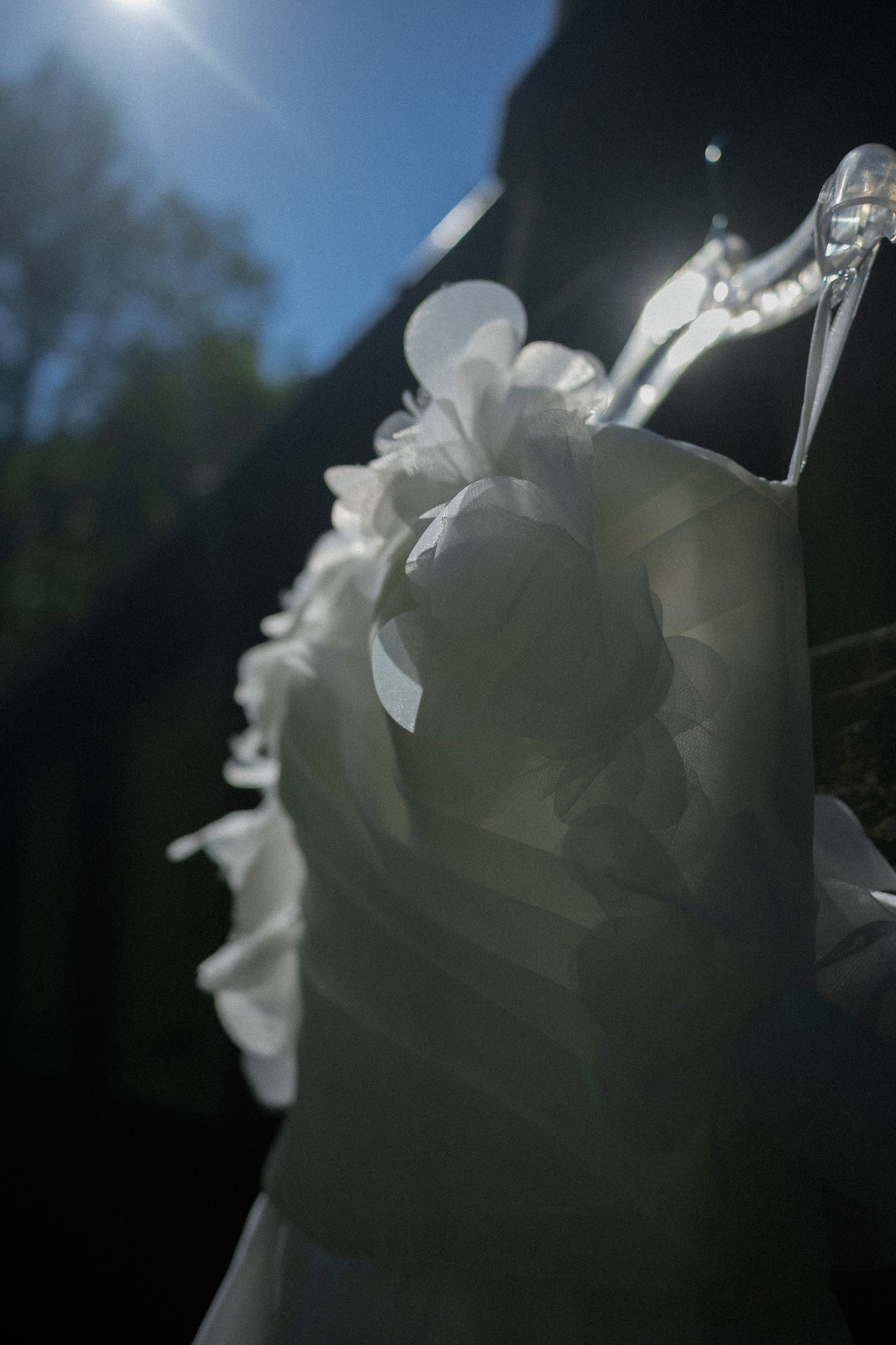 White wedding dress with floral details hanging on a clear hanger, backlit by sunlight against a dark background.