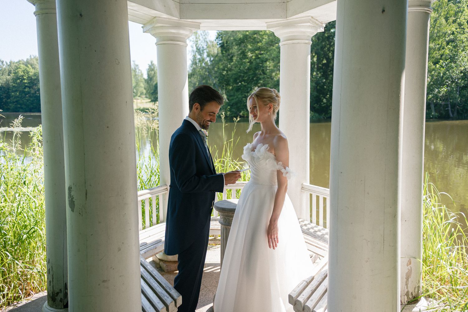 Bride and groom exchange vows in elegant white gazebo by serene lake, surrounded by lush greenery on their wedding day.