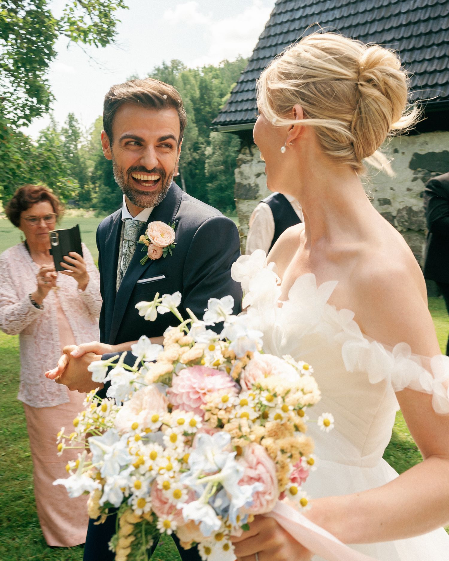 Smiling groom in navy suit holds hands with bride in lace dress holding floral bouquet at outdoor wedding ceremony.