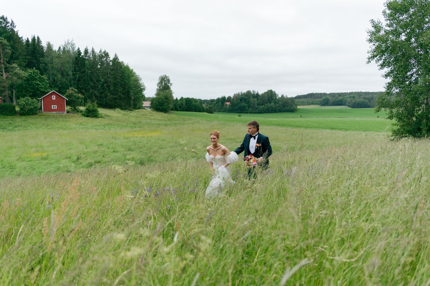 Wedding party walking through a lush green meadow with forest and red barn in background.