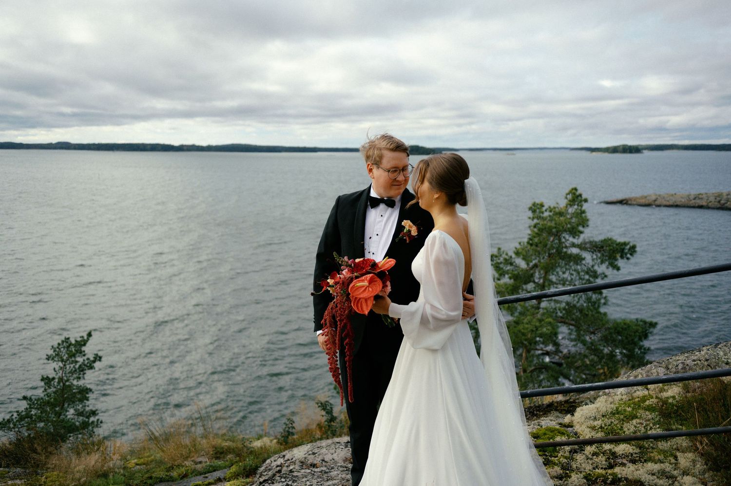 A couple in formal wedding attire shares a romantic moment by a scenic waterfront under cloudy skies.