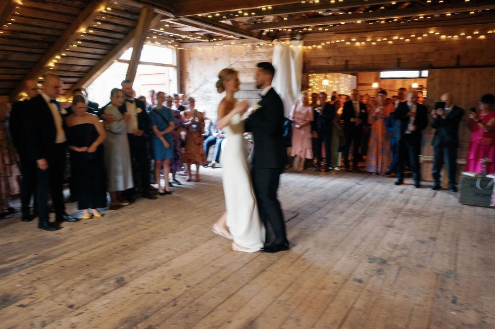 Couple shares their first dance surrounded by guests in a barn venue with twinkling lights.