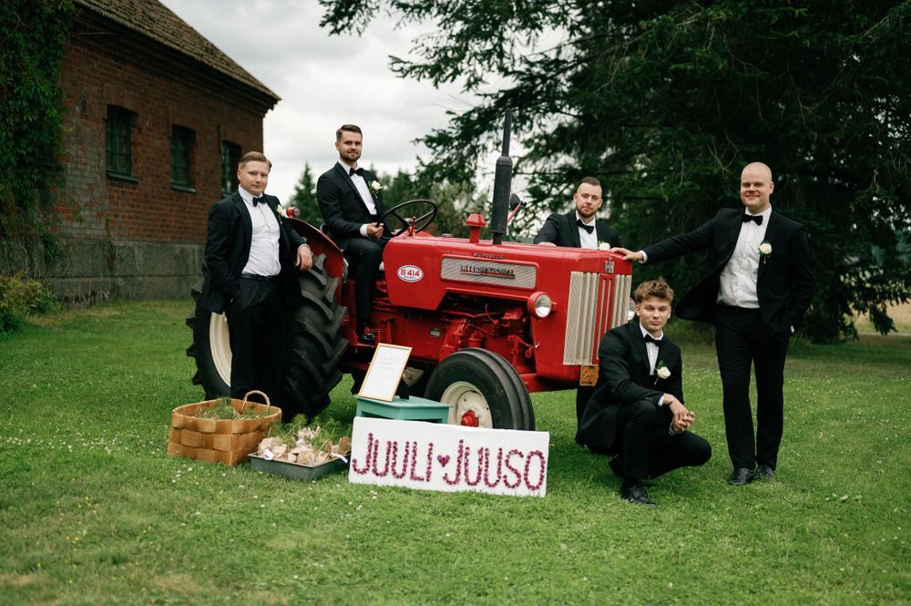 Wedding party posing outdoors with a vintage red tractor against a brick building backdrop.
