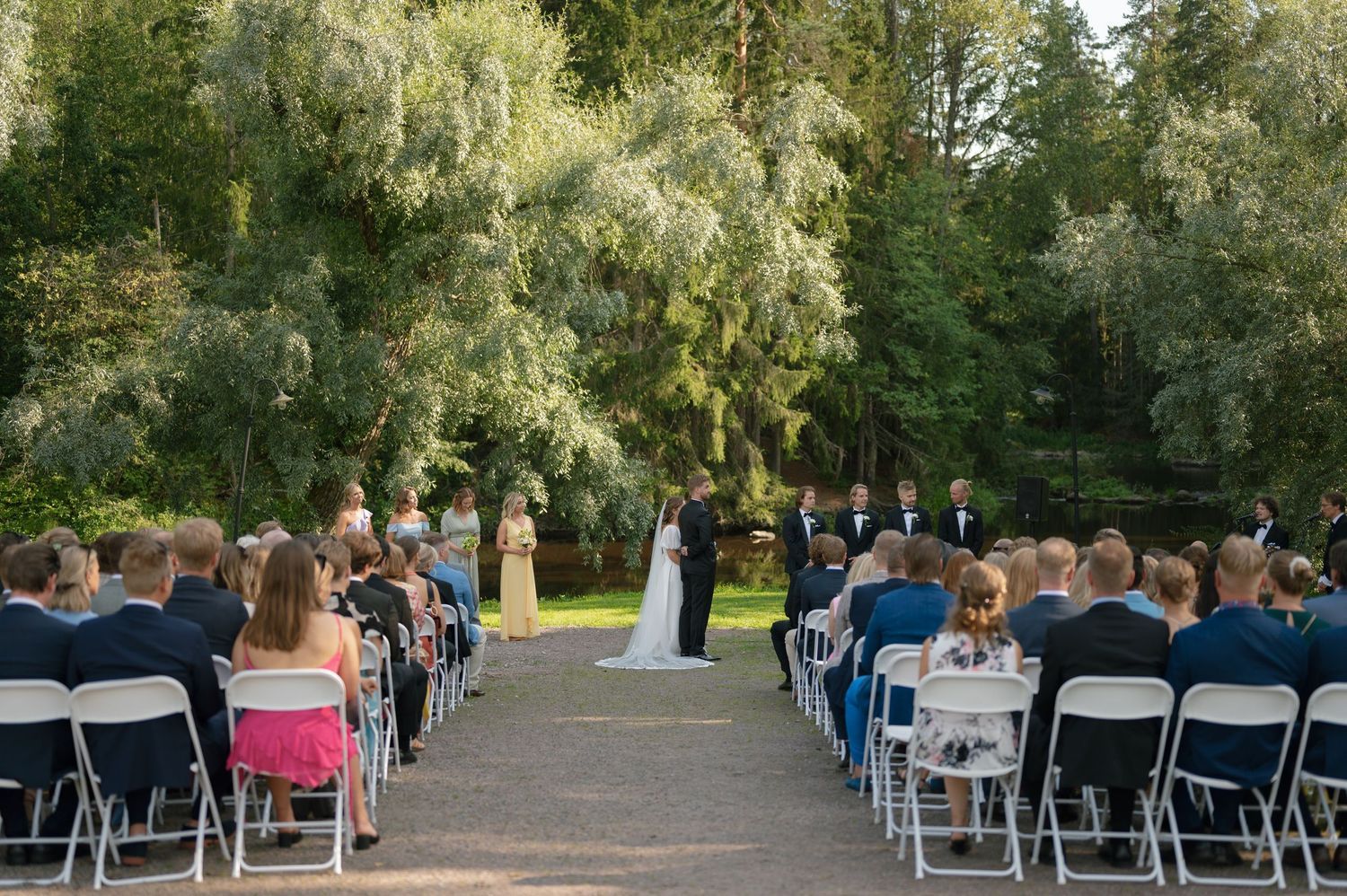 Outdoor wedding ceremony setup with rows of white chairs on a lawn surrounded by tall trees.