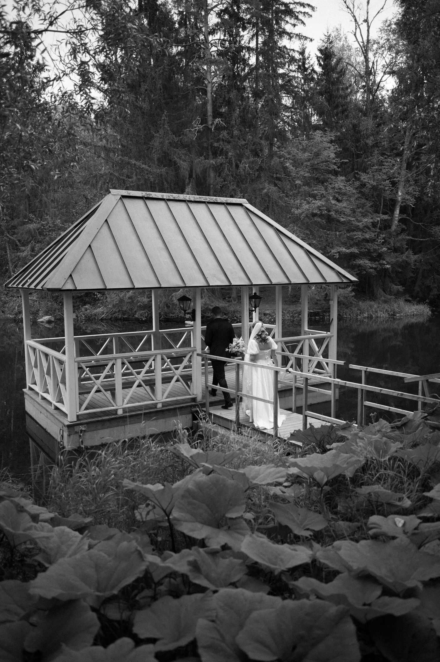 A black and white wedding portrait in a rustic gazebo surrounded by large leafy plants.