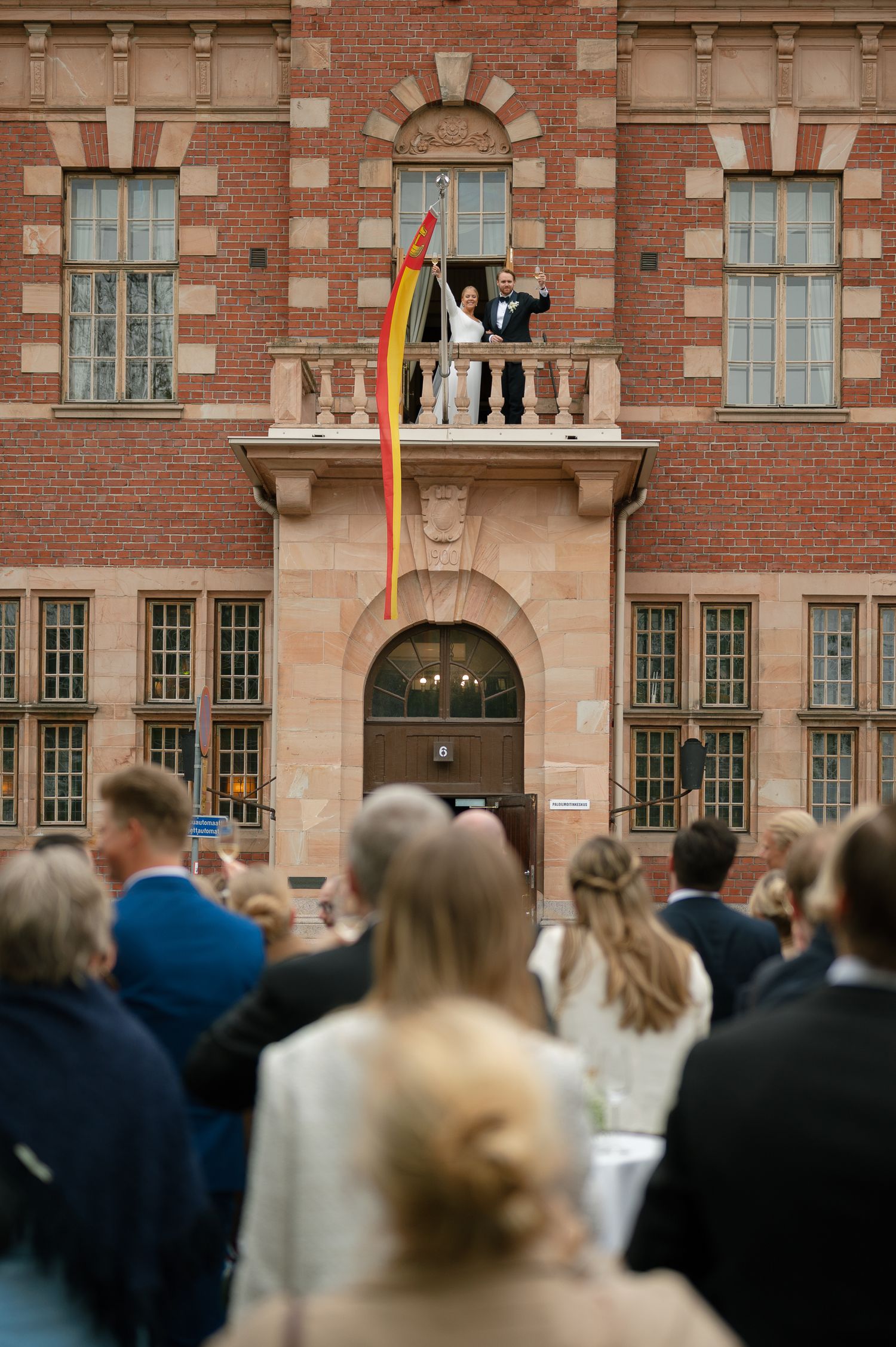 A crowd gathers outside a historic brick building with ornate windows and arched details during a ceremony.