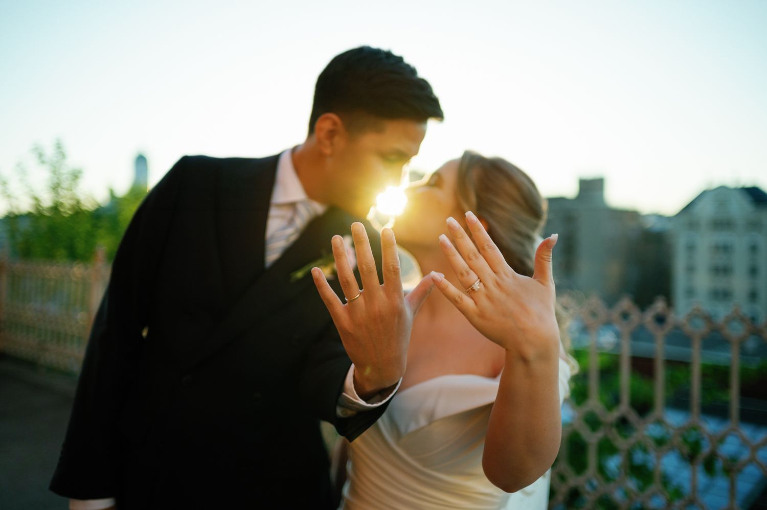 Newlywed couple kissing at sunset, showing off wedding rings to camera outdoors.