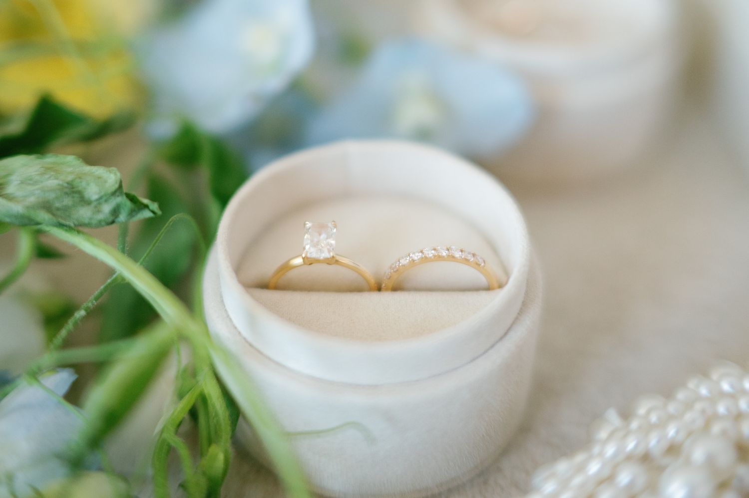 Two gold wedding rings, one solitaire diamond engagement ring, displayed in a white velvet ring box with flowers.