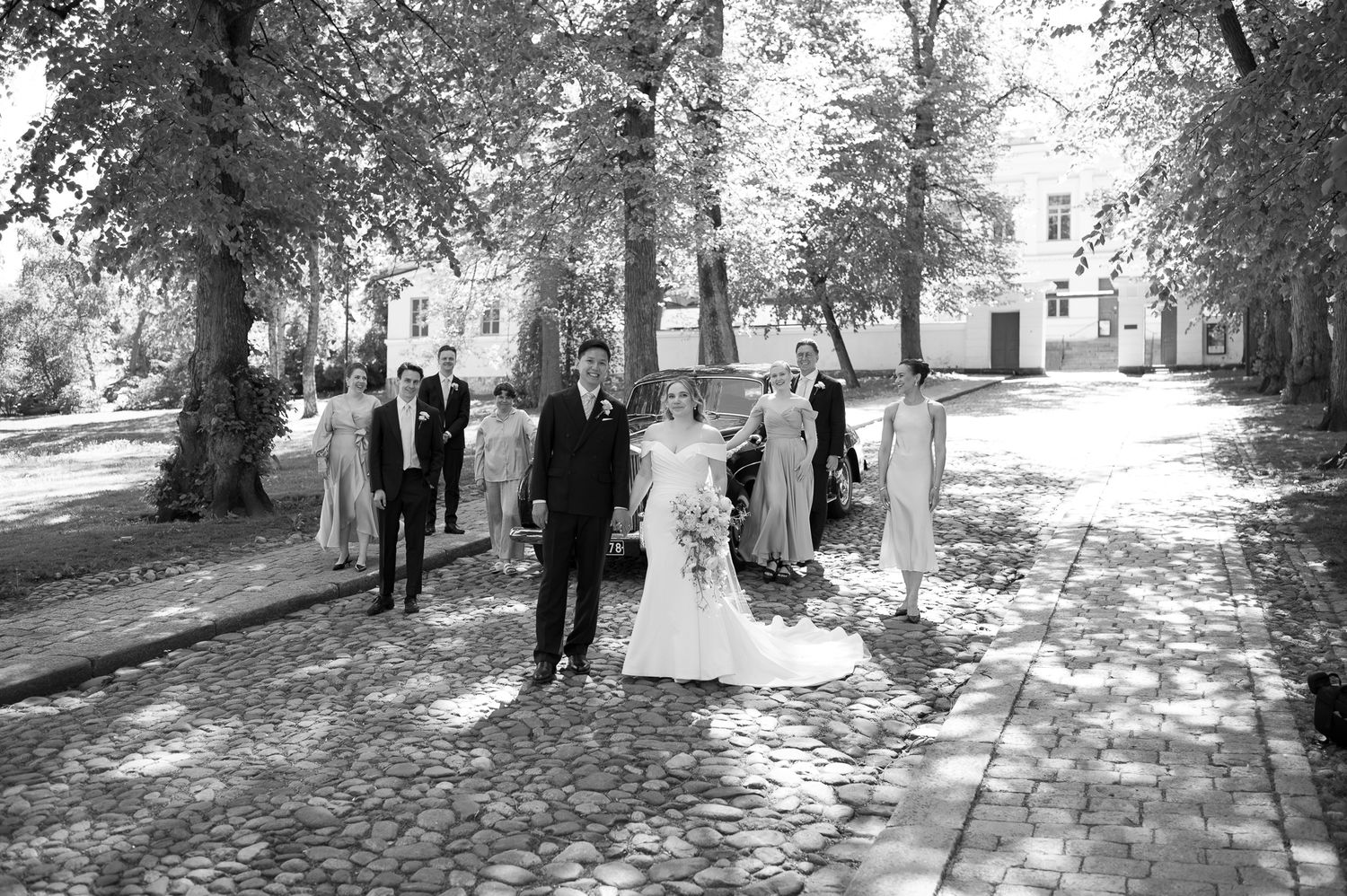 Black and white wedding photo of bride and groom leading bridal party along tree-lined cobblestone path.