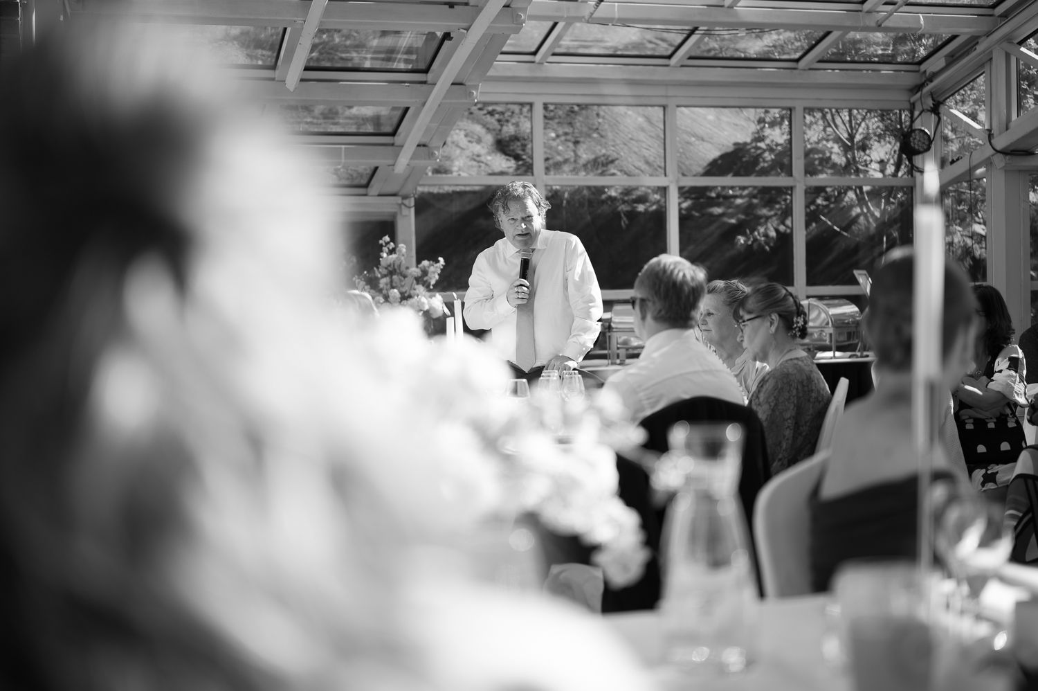 Black and white photo of a man giving a speech at an elegant indoor wedding reception dinner.
