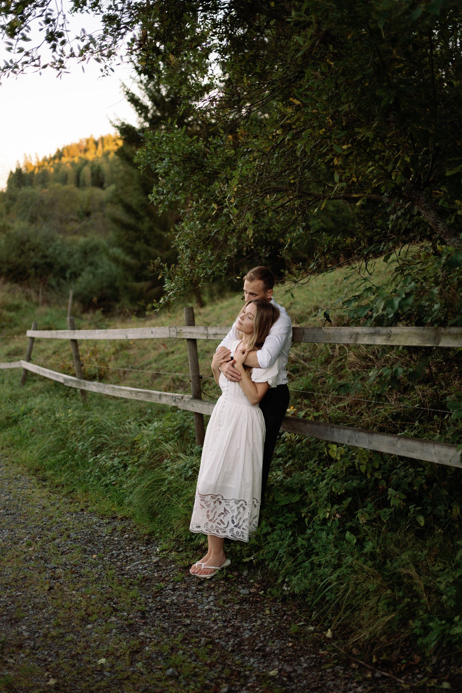Couple embracing by a rustic wooden fence surrounded by lush green trees during golden hour outdoors.