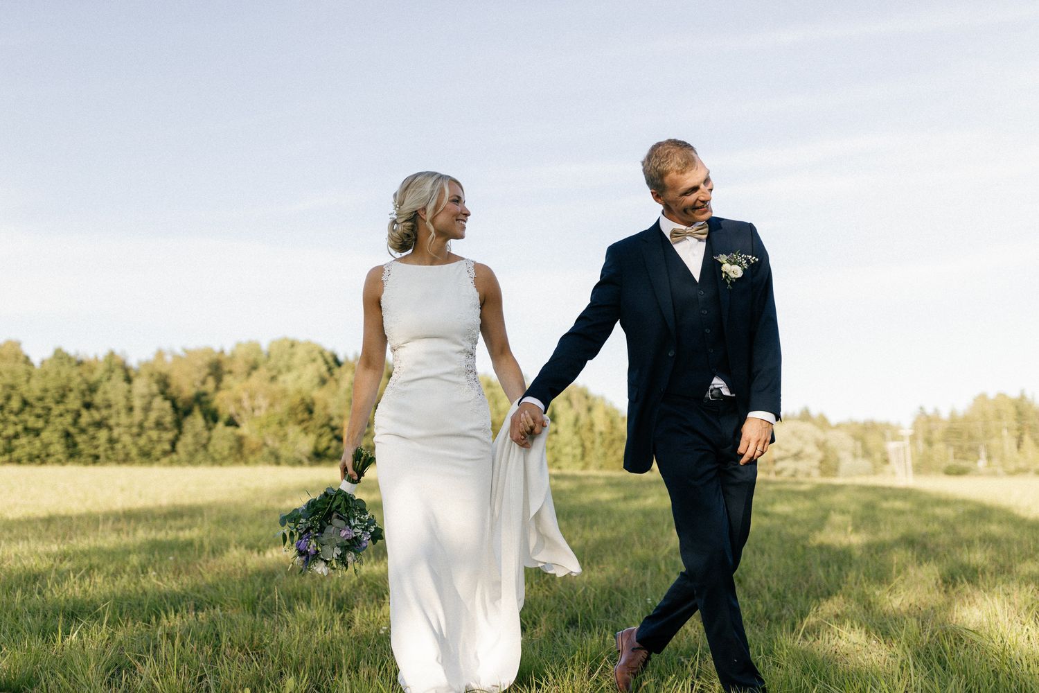 Elegant couple walking hand in hand through sunny meadow in wedding attire.