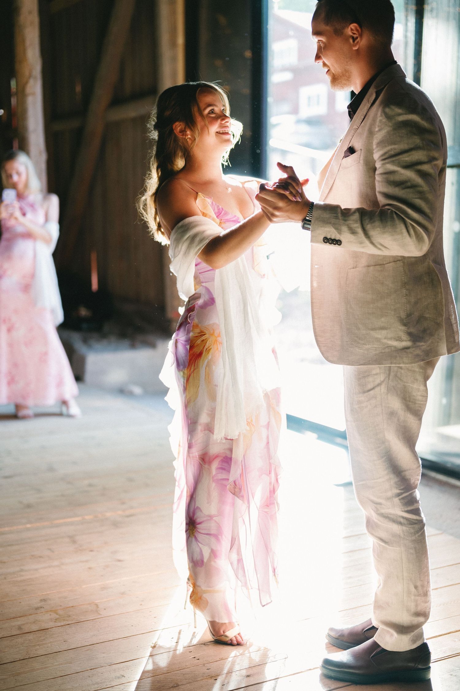 A couple dances together in a sunlit rustic venue, with the person in pink floral dress smiling brightly.