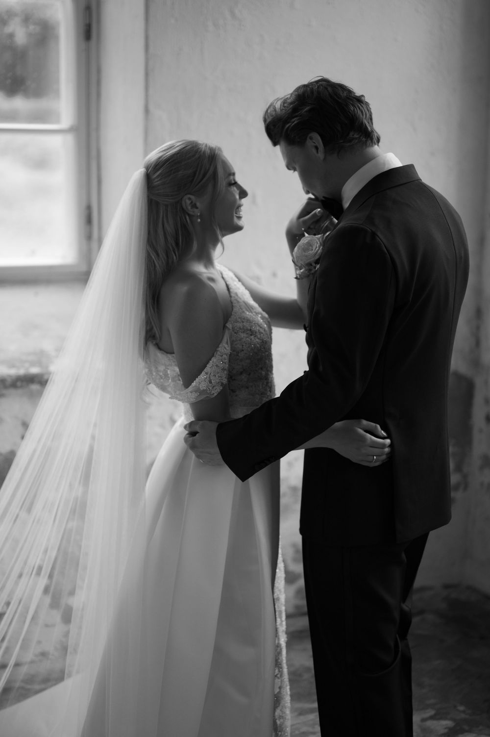 An intimate black and white wedding photo showing the couple in a gentle embrace by a window.