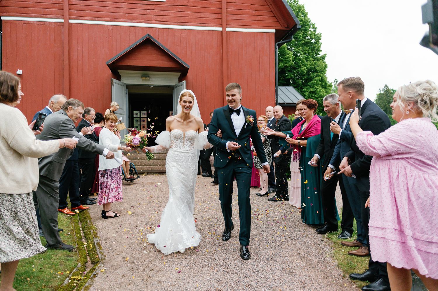Wedding party exits red church building while guests line pathway.