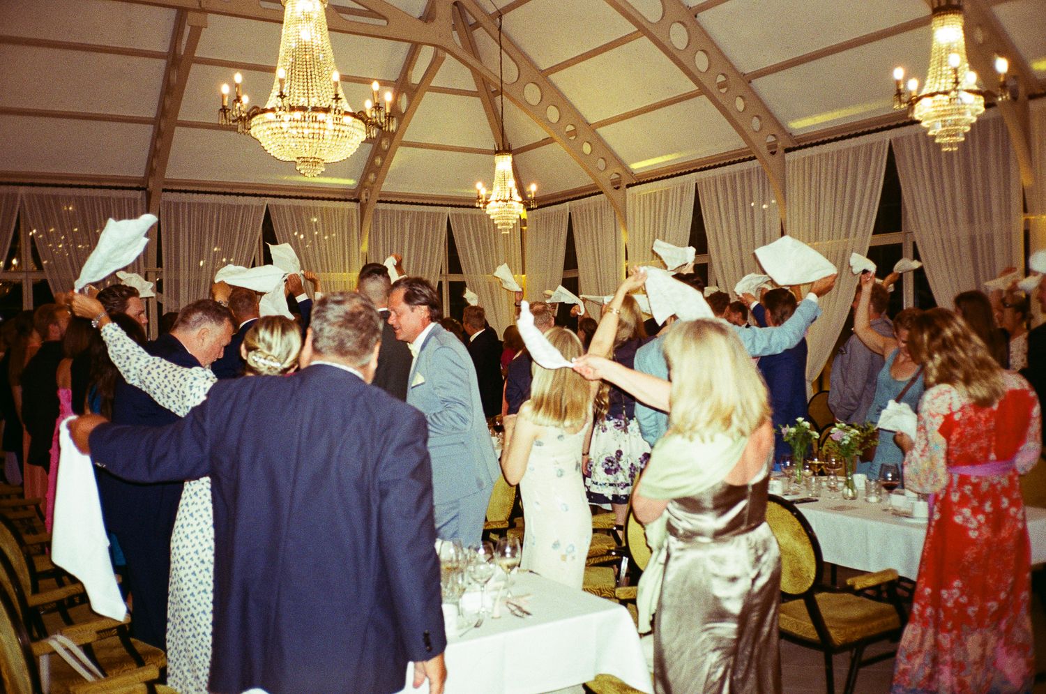 Guests wave white napkins at a festive wedding reception in an elegant tent venue with chandeliers.