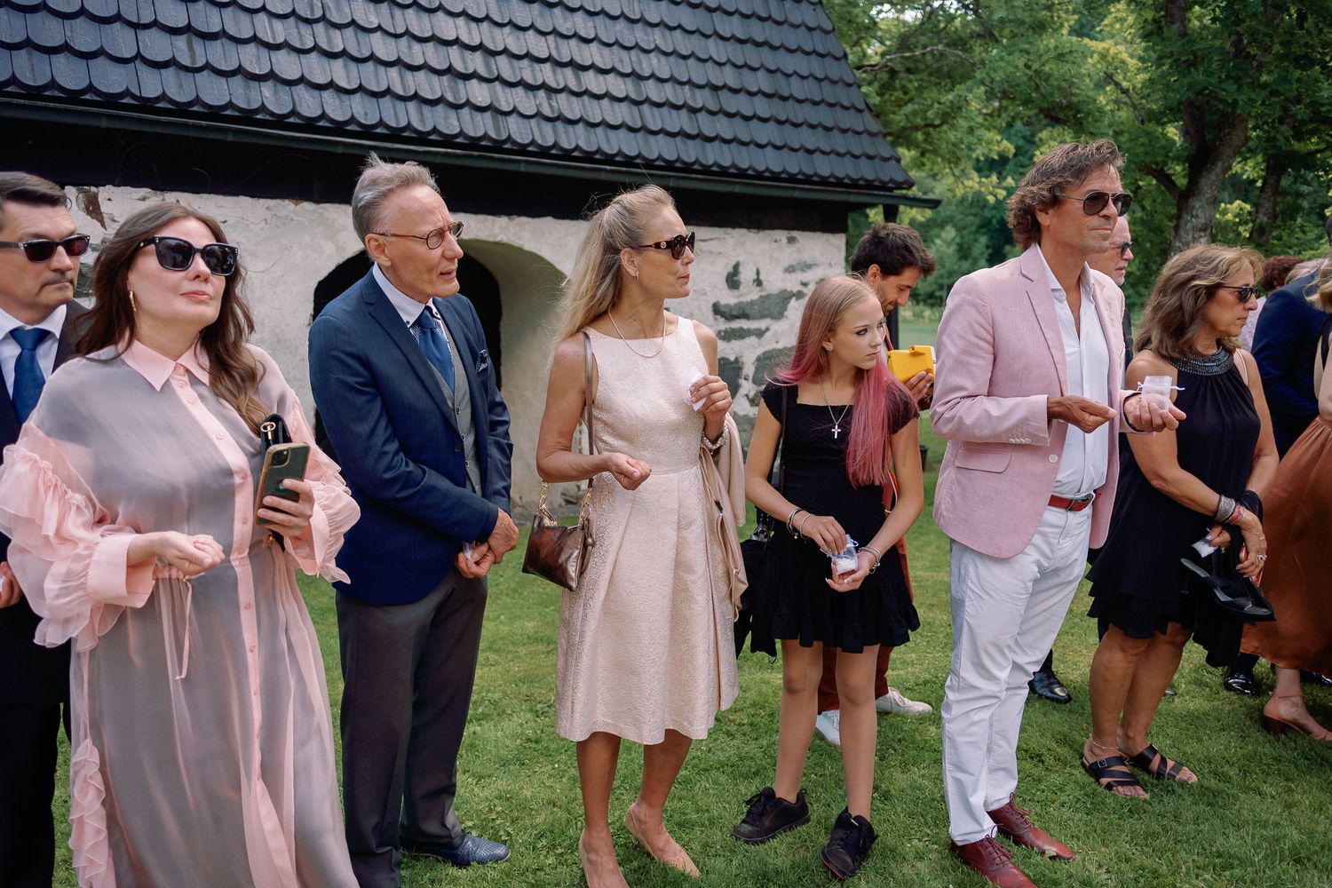 Elegantly dressed guests gather outdoors at a summer wedding ceremony, including women in pink dresses and a man in a pink blazer.