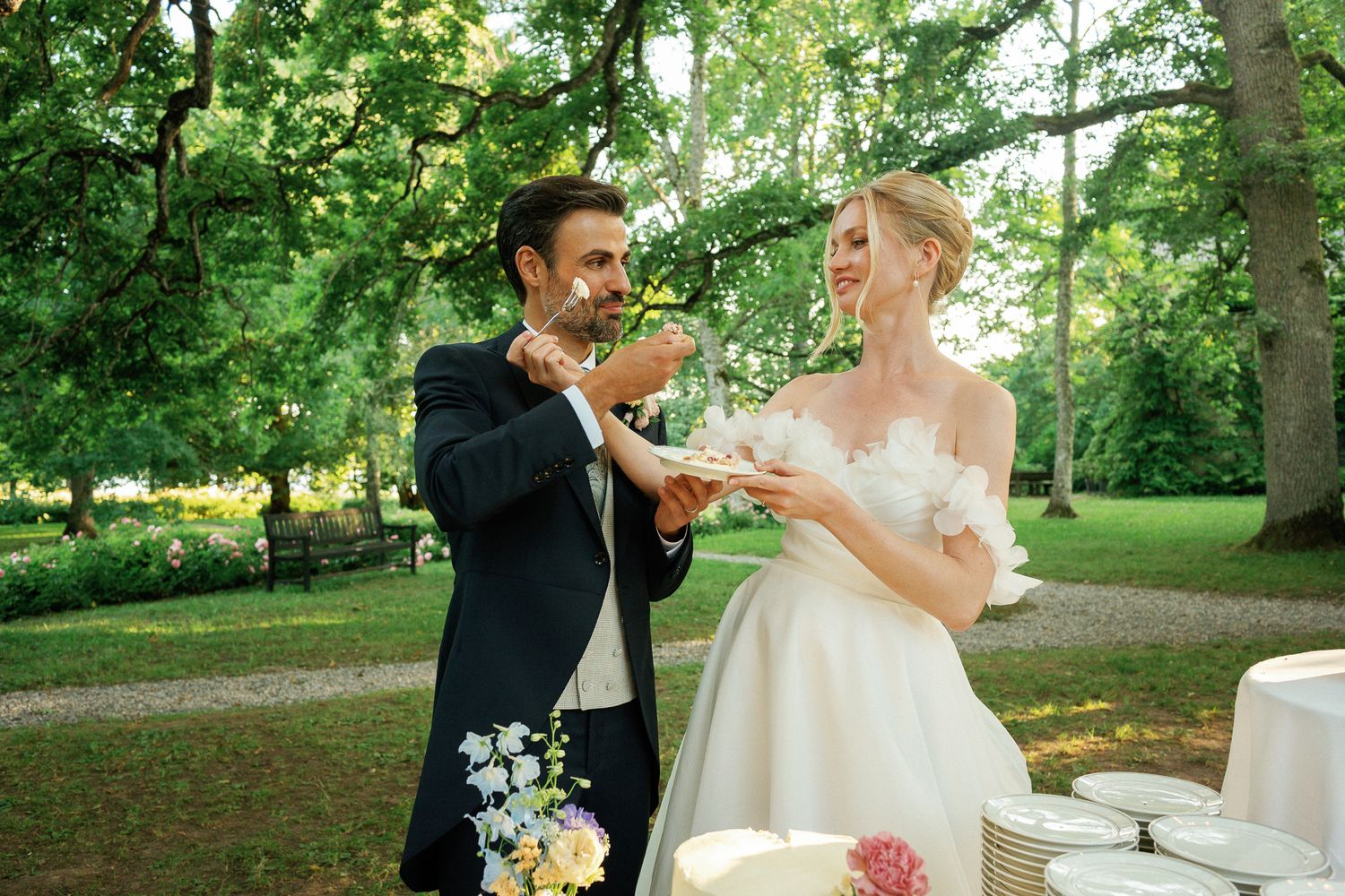 Bride and groom cutting wedding cake outdoors in lush green garden, bride wearing off-shoulder white gown.