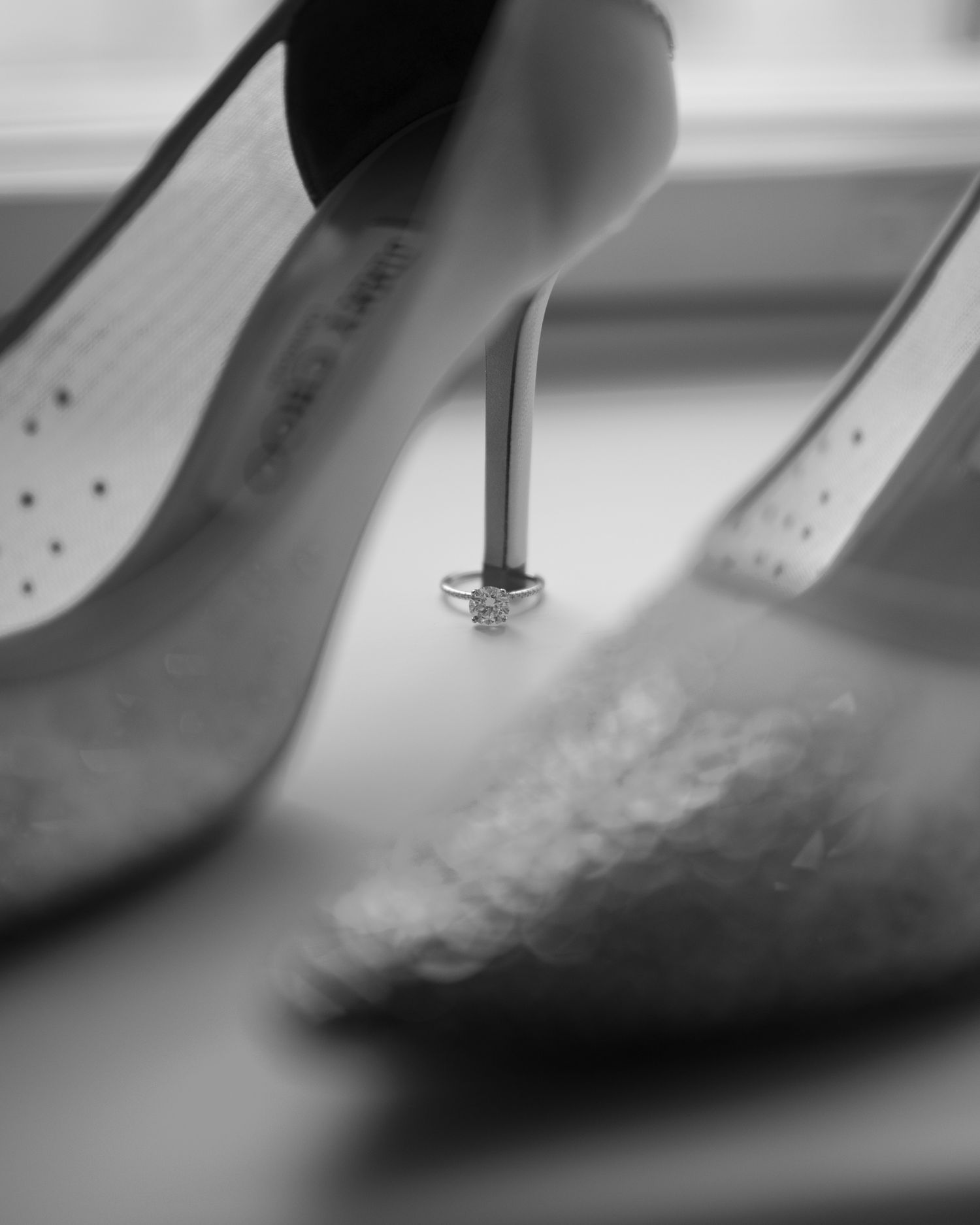 Black and white photo of elegant high heels with a diamond engagement ring balanced between the stiletto heels.