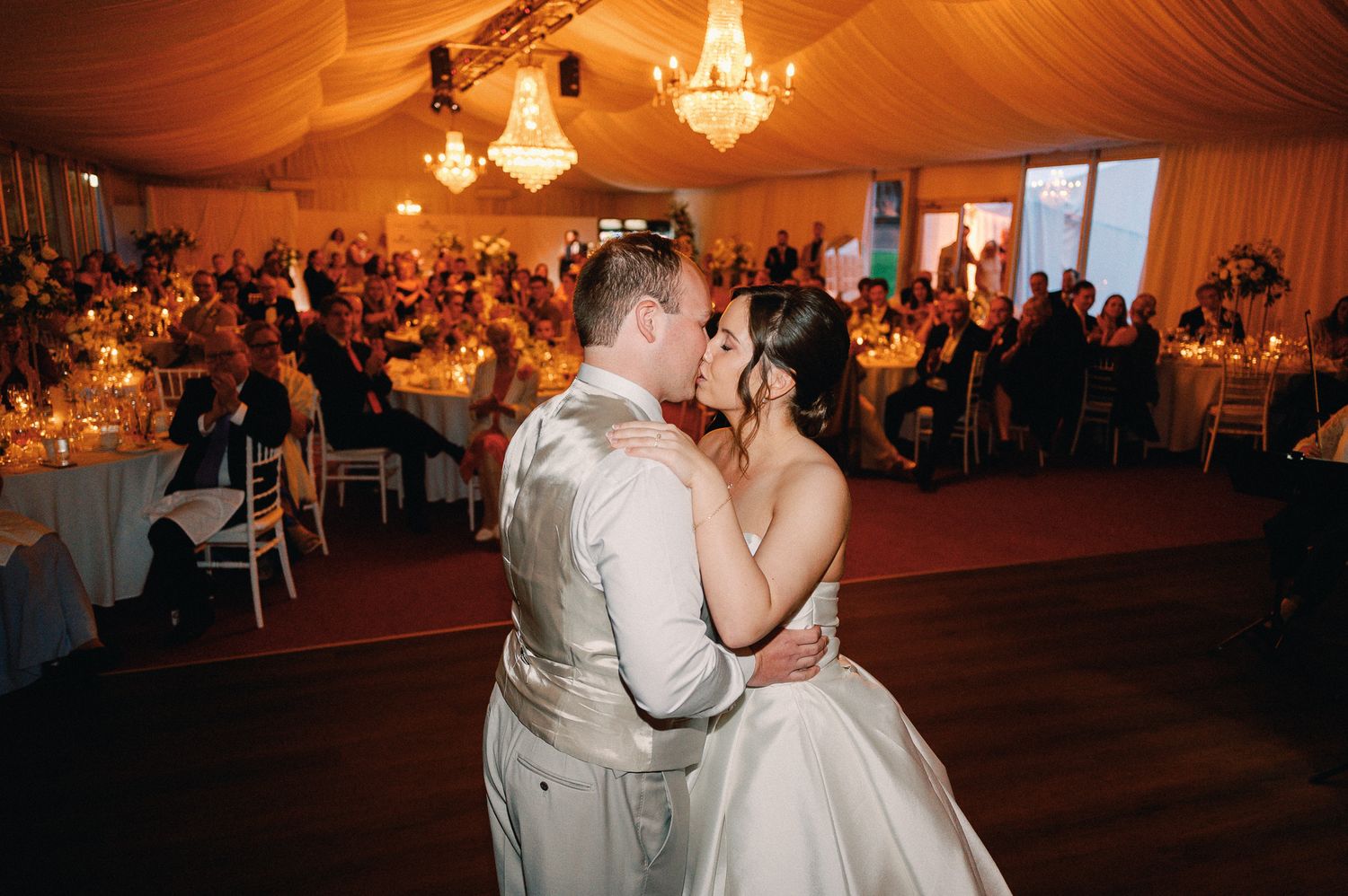 Bride and groom share first dance kiss in elegant candlelit wedding reception tent with chandeliers and guests seated.