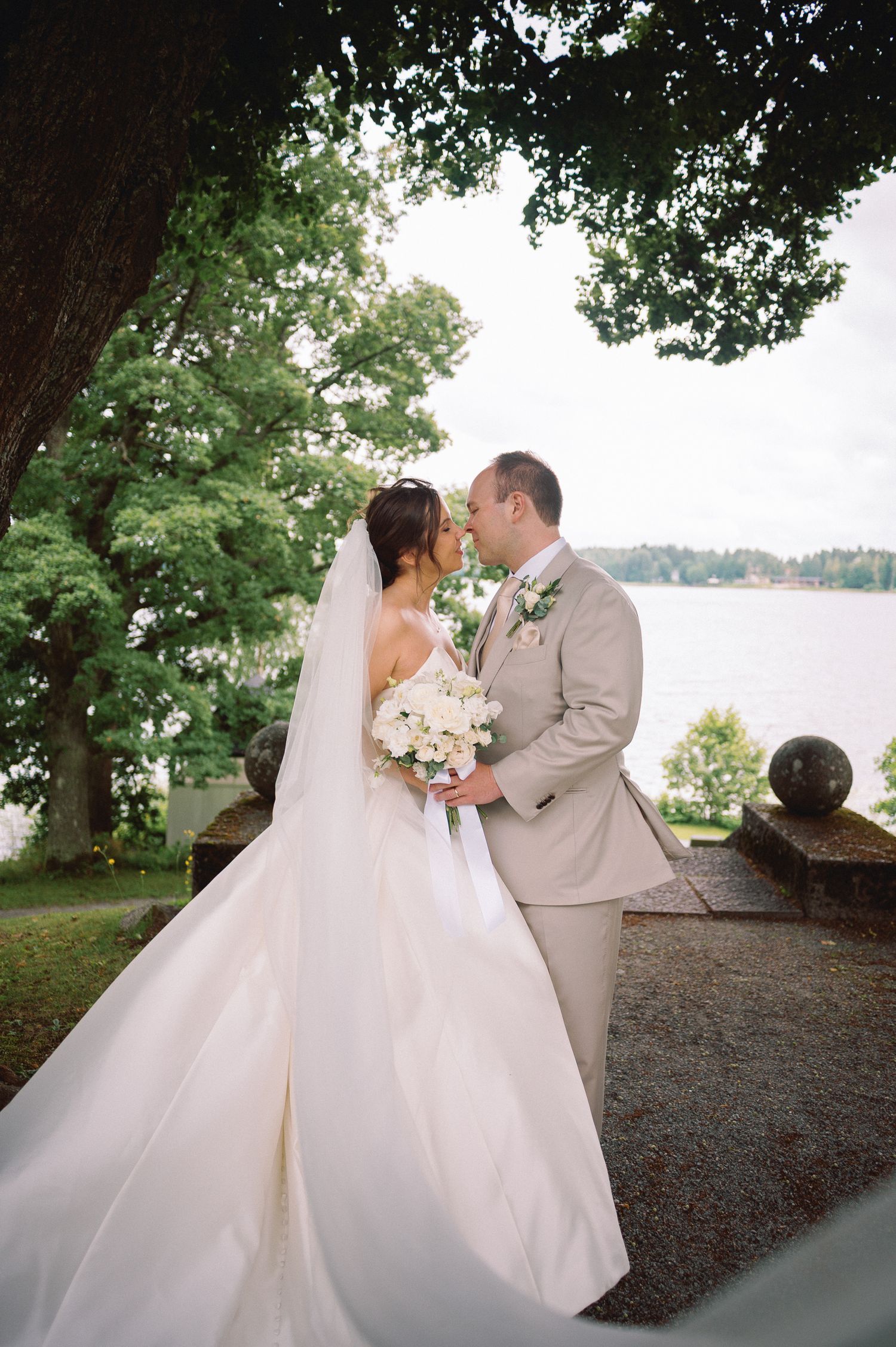 Bride and groom share a romantic moment by a lakeside, bride in elegant white gown with veil holding white bouquet.