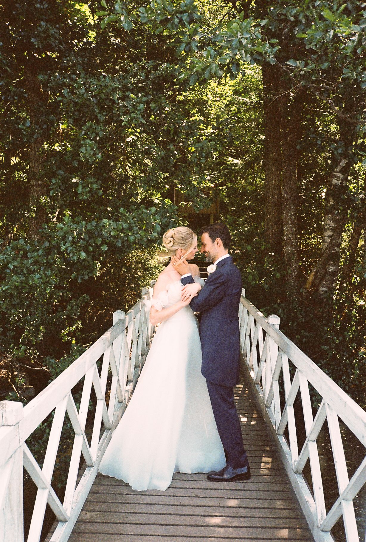 Bride and groom share a romantic moment on a white wooden bridge surrounded by lush green trees.