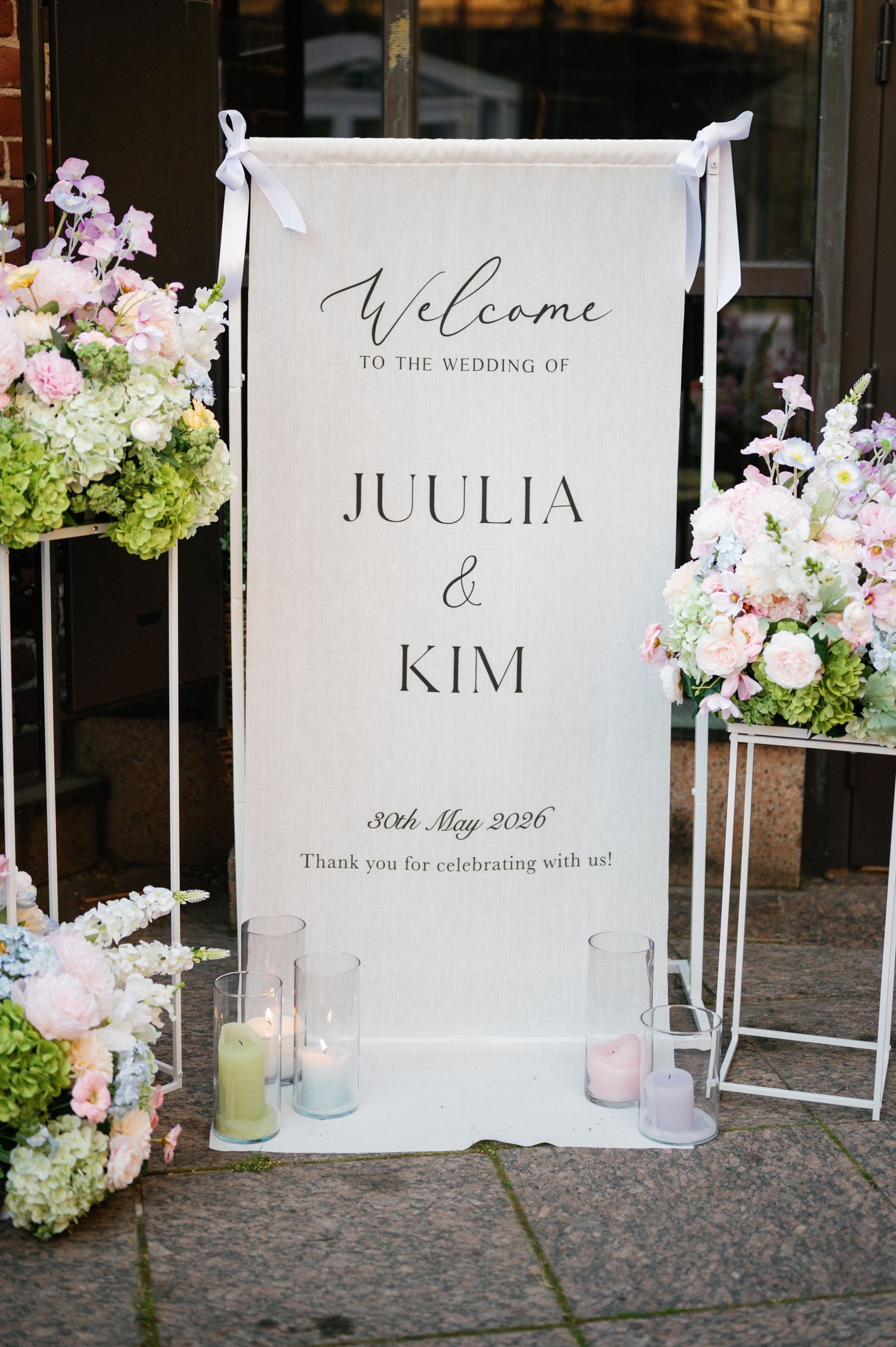 Elegant wedding welcome sign for Juulia and Kim, May 30 2026, flanked by pastel floral arrangements and candles.