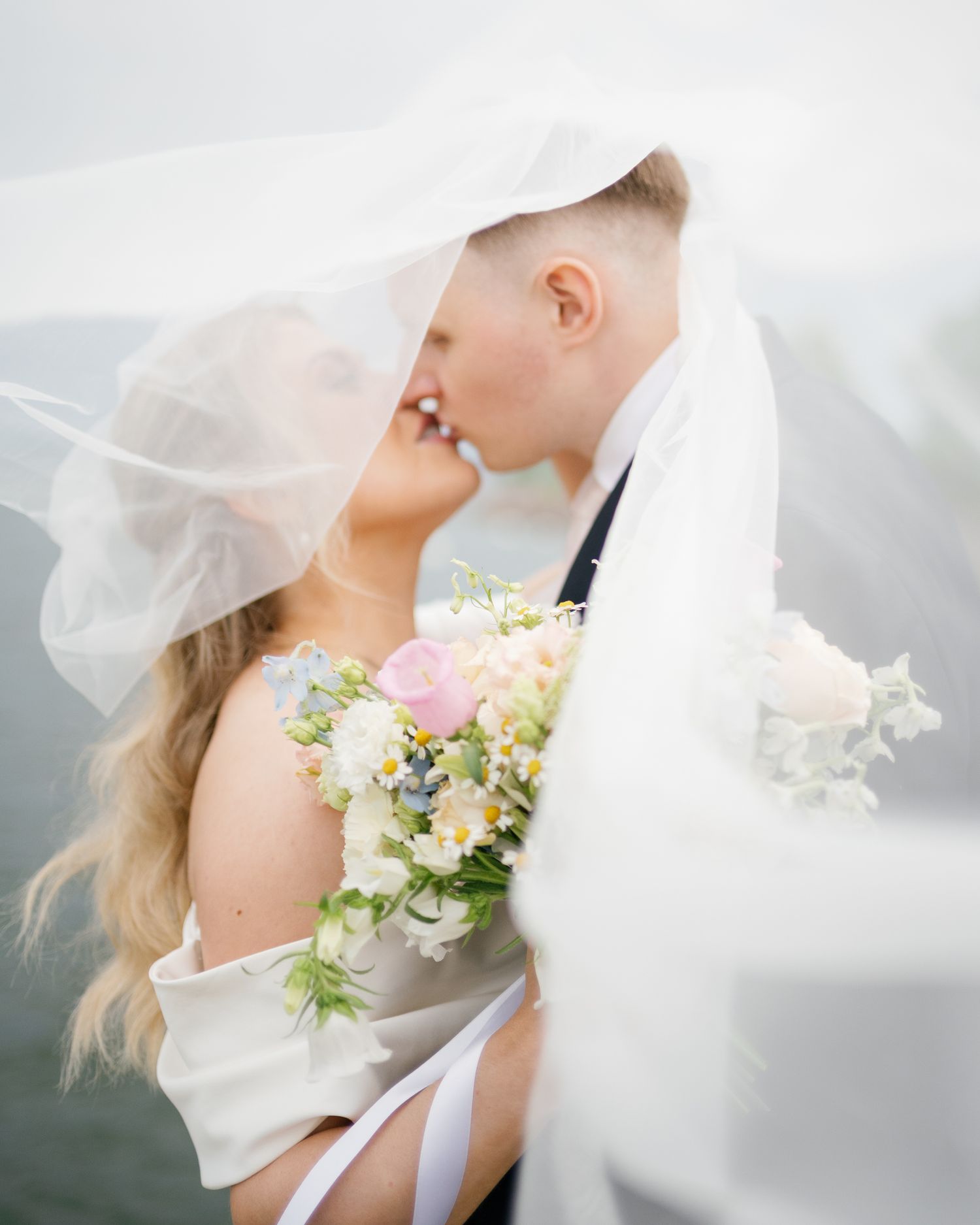 Bride and groom kissing under flowing white veil, bride holding pastel spring bouquet with pink roses and wildflowers.