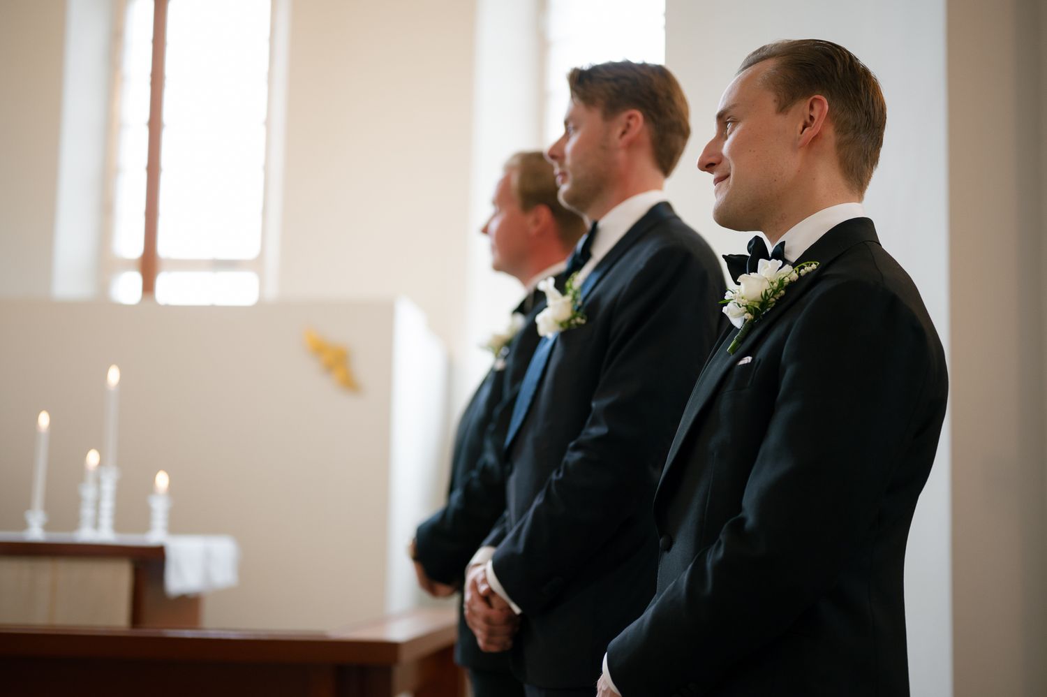 Three groomsmen in dark suits stand at attention during a church wedding ceremony.