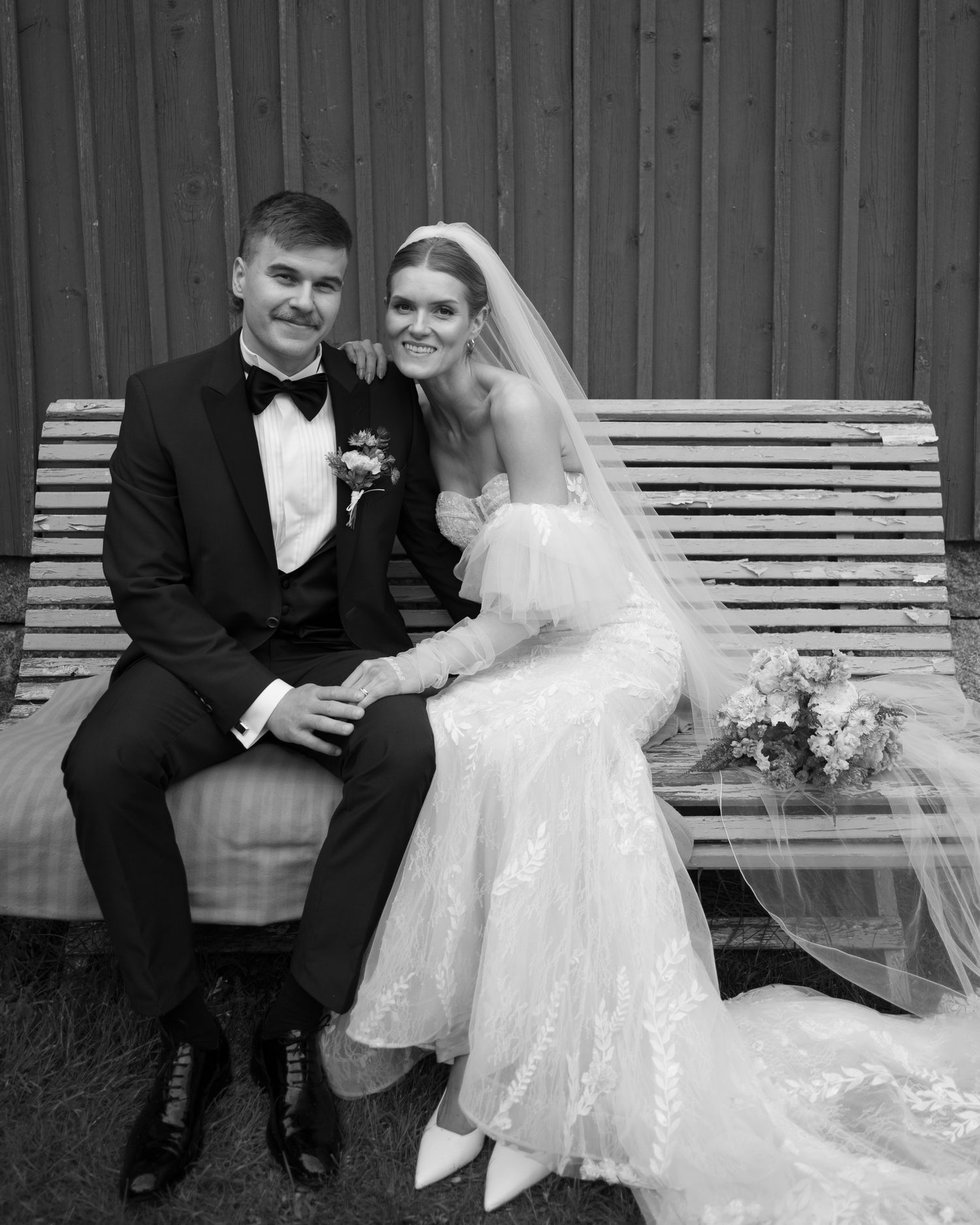Black and white wedding photo of couple sitting on white bench against wooden wall.