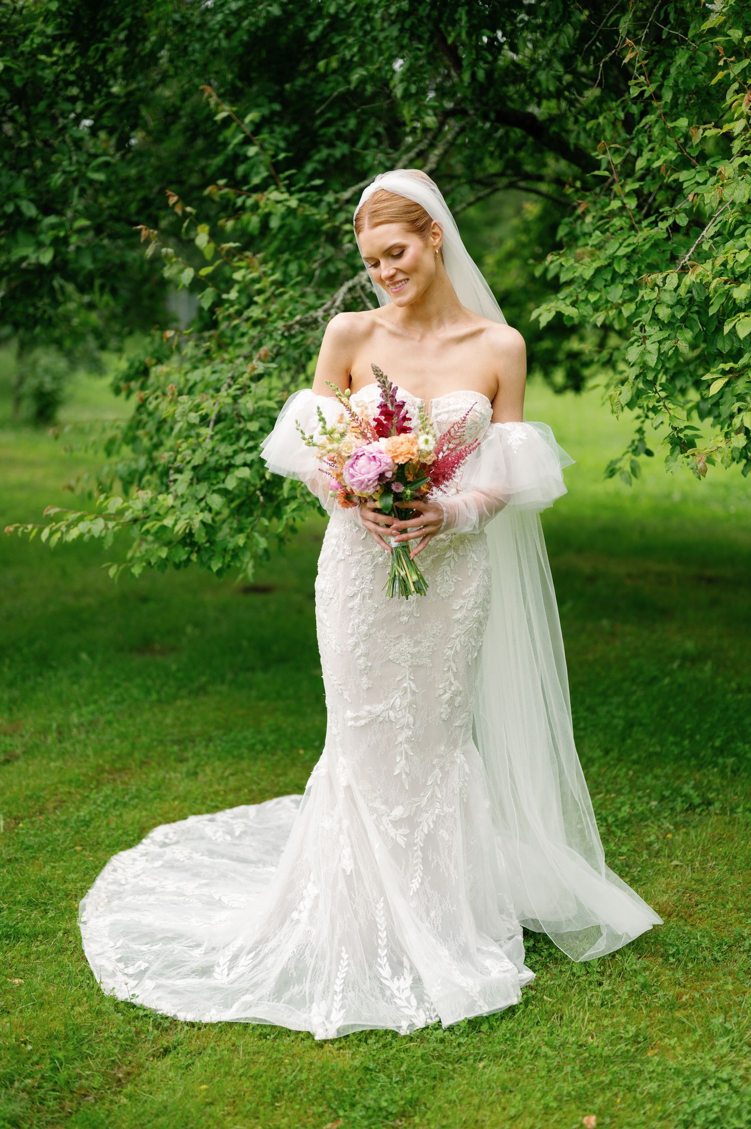 Bride in a detailed lace wedding gown poses with her bouquet in a lush garden setting.