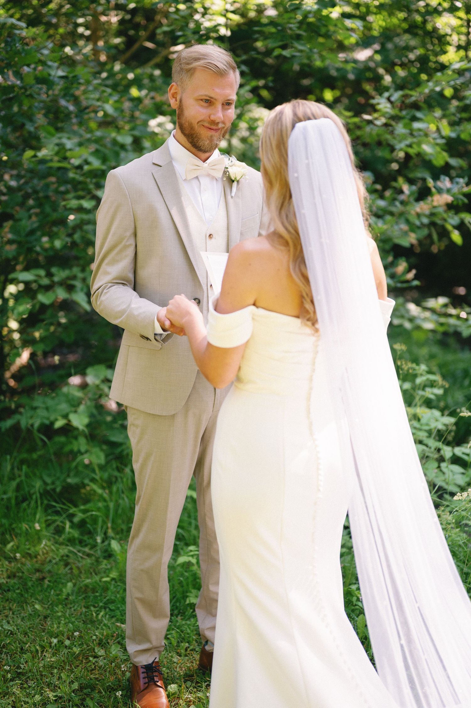 A bride in a flowing white dress and groom in beige suit read letters to each other in a garden setting.