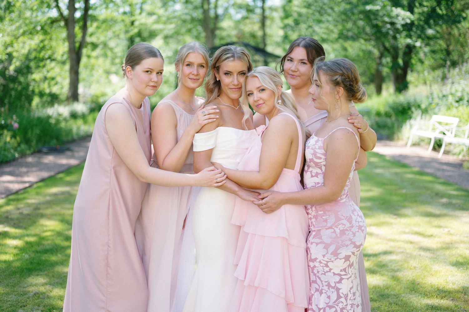 Wedding party in pink and white dresses pose together in a sunny garden setting.