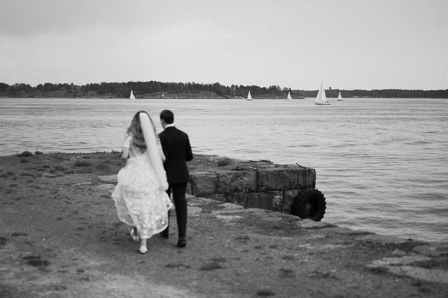 Black and white photo of a couple by the water with sailboats in the distance.